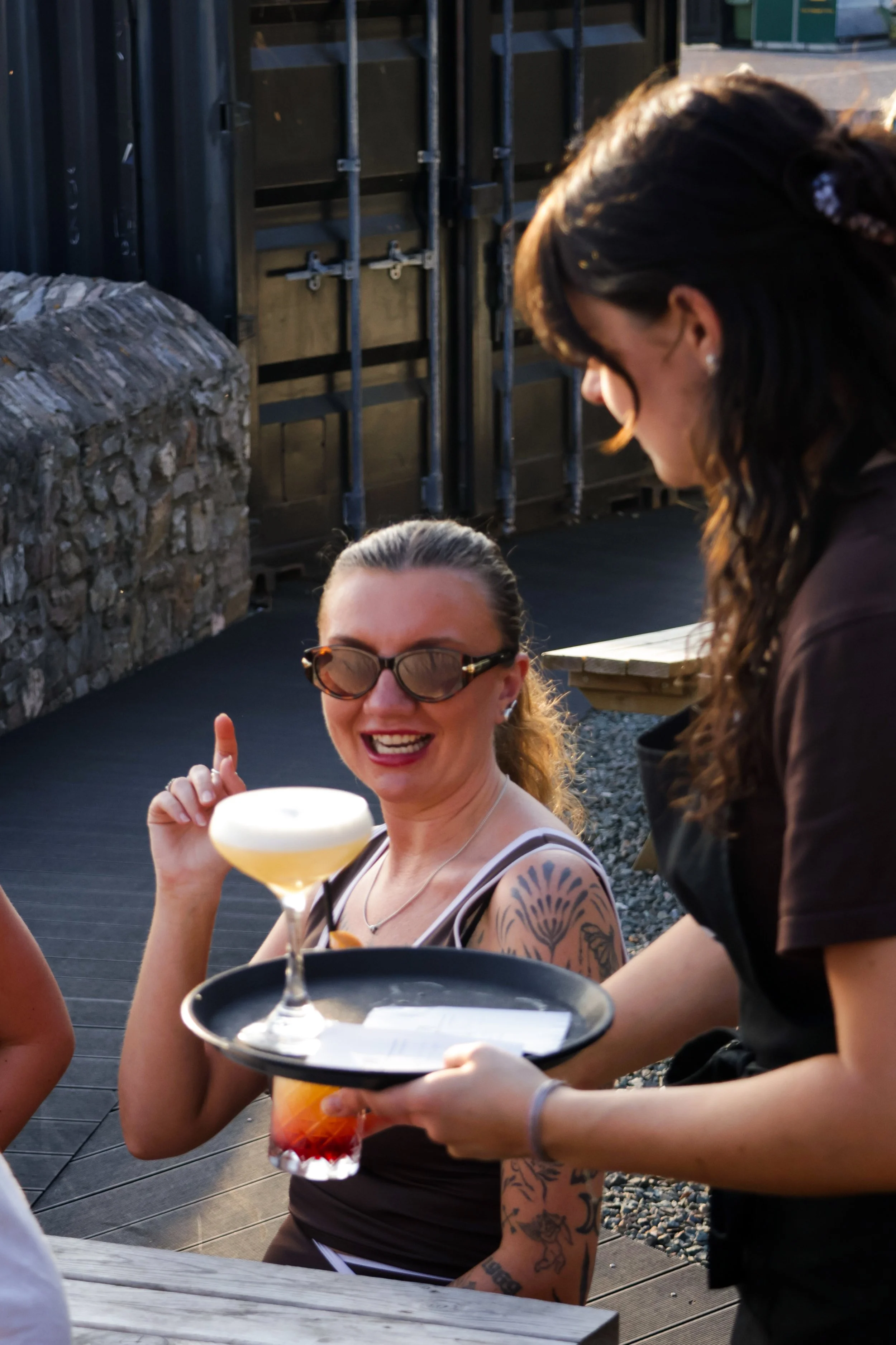 A woman with glasses and tattoos smiling and holding a cocktail glass, sitting at an outdoor table with a server presenting a drink tray