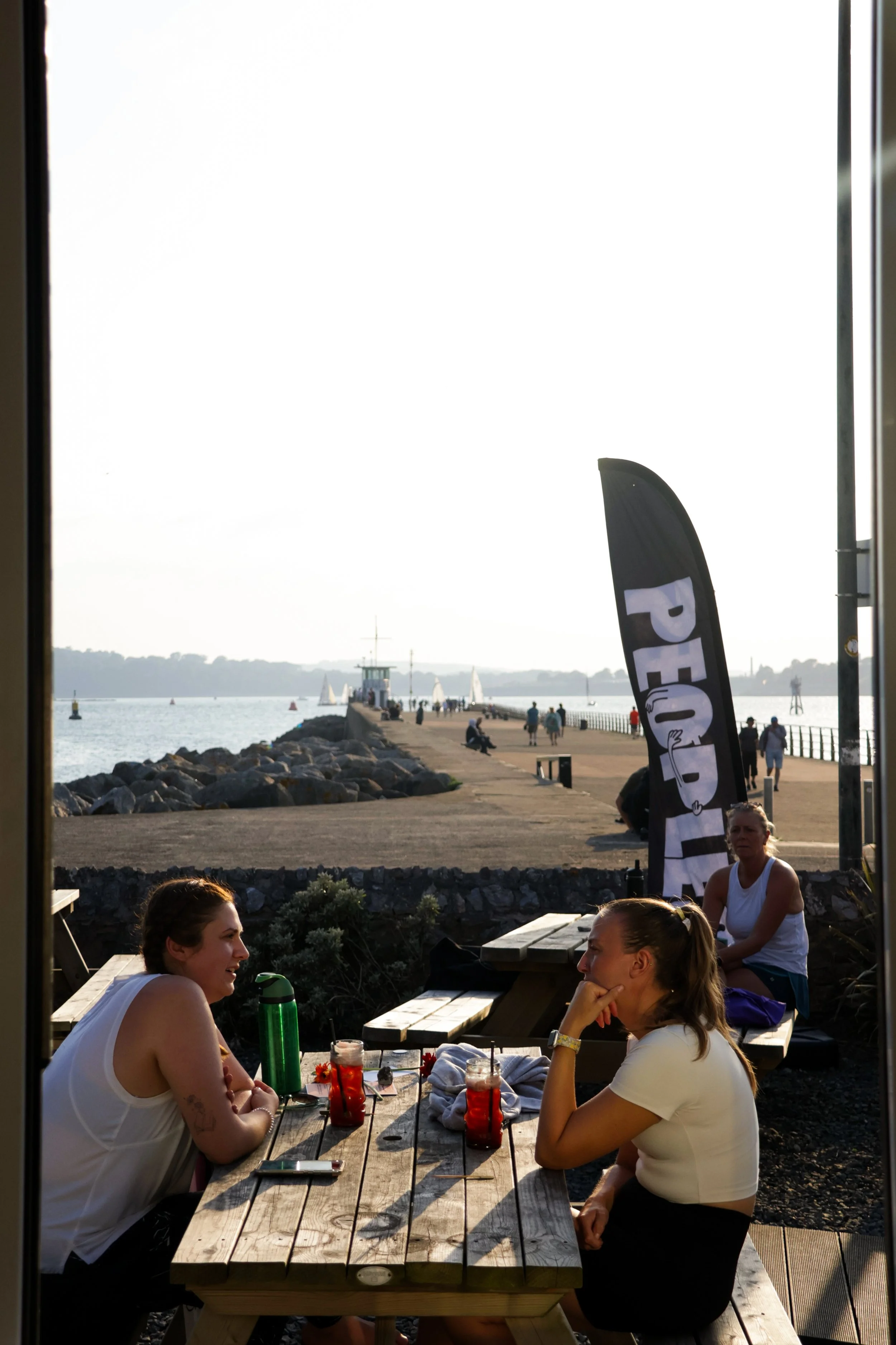 People sitting at a wooden table outdoors near a waterfront, with sailboats and a pier visible in the background, and two women smiling and talking.