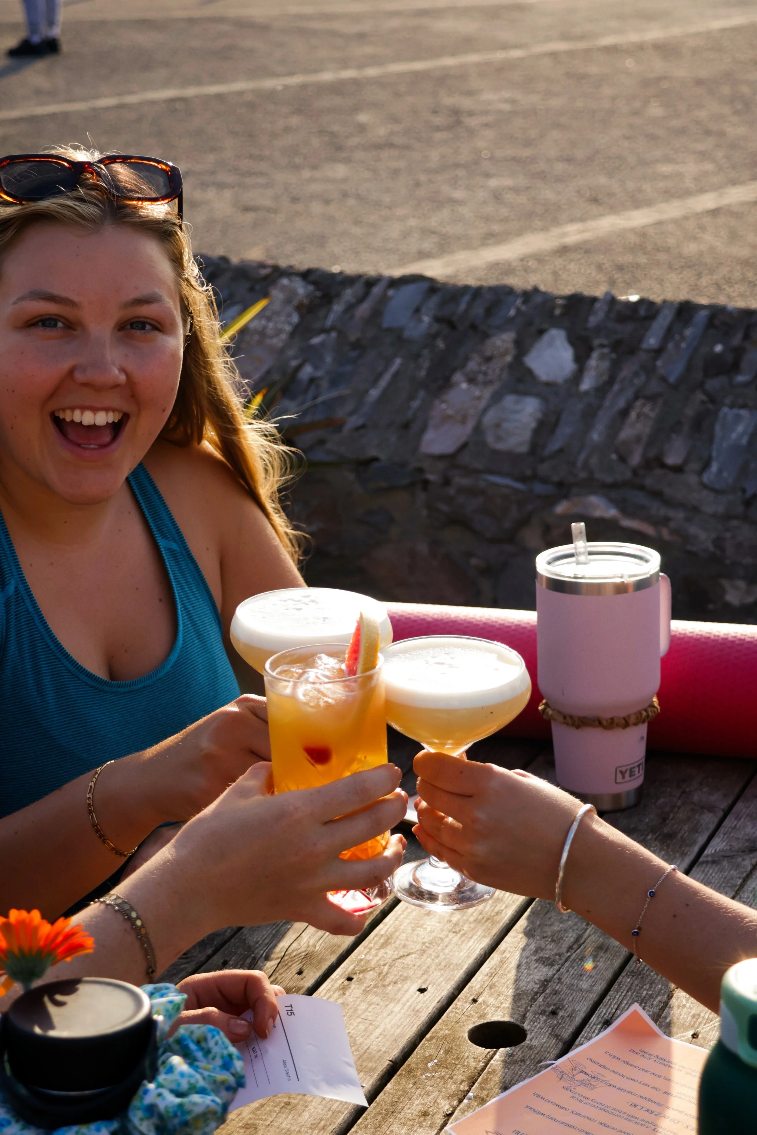Two women clinking drinks at an outdoor table, with one woman smiling widely, wearing a blue tank top, sunglasses on her head, and jewelry, while holding a cocktail with a citrus garnish. The table has a pink tumbler, papers, and a small plant, with 