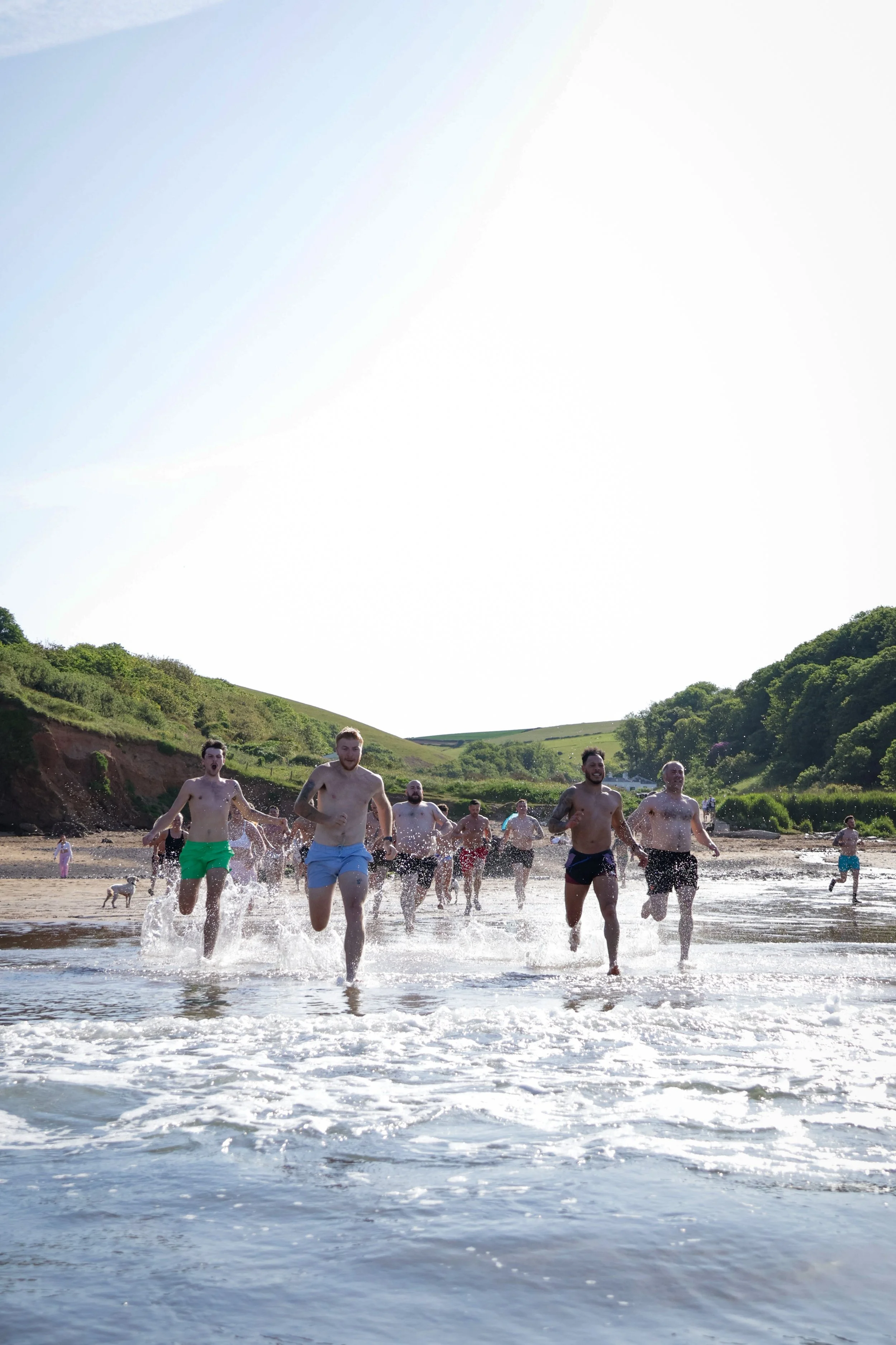 A group of men and women running into the ocean at the beach, with green hills and trees in the background.