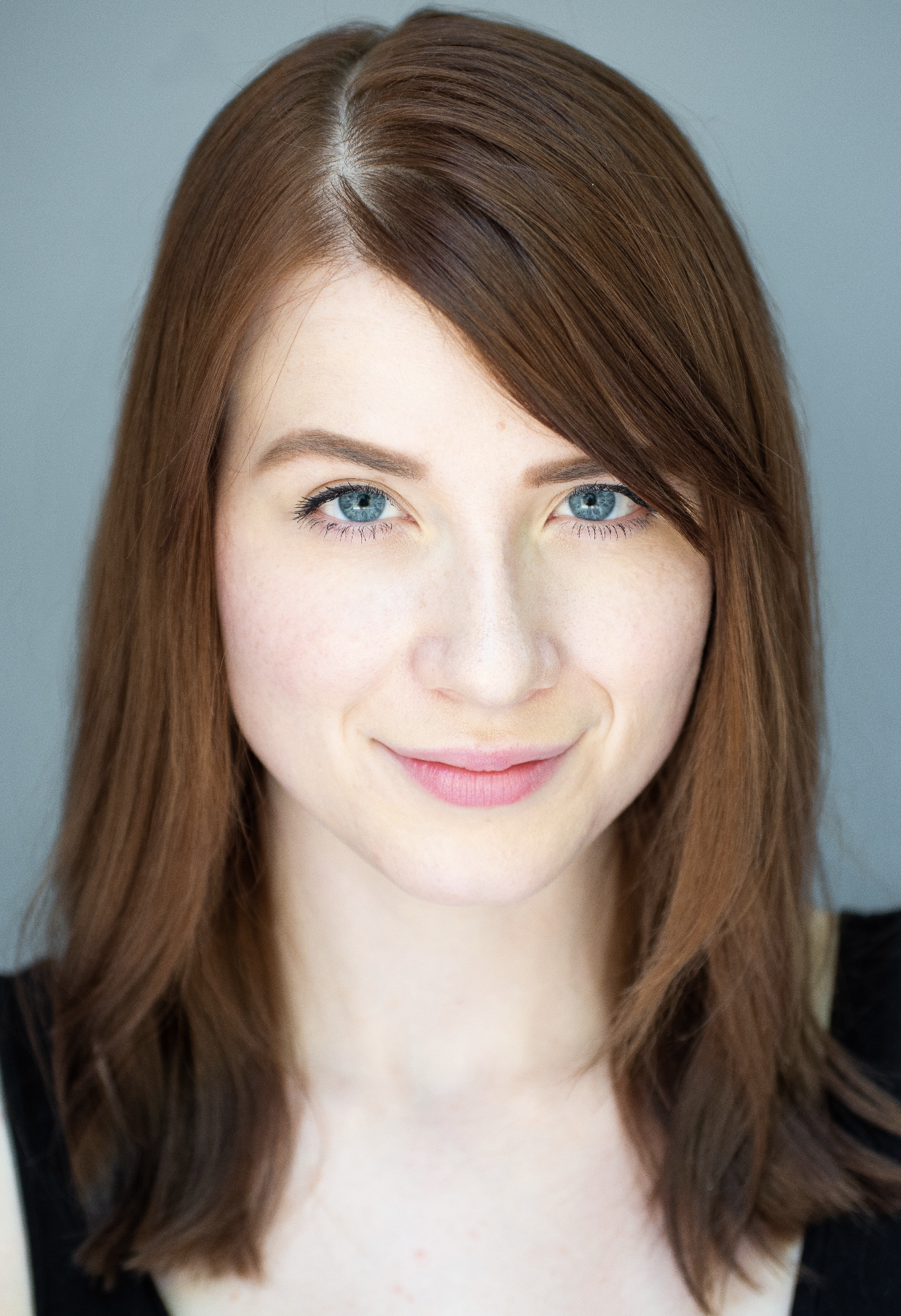Close-up of a young woman with long auburn hair, blue eyes, and a slight smile, wearing a black top, against a gray background.