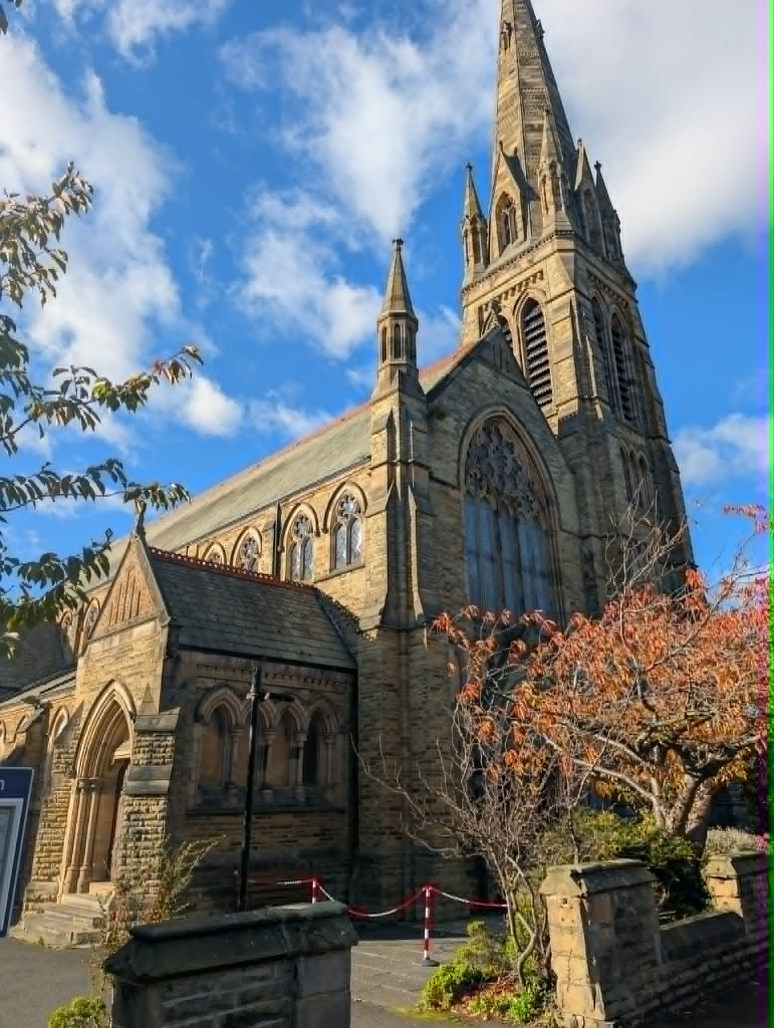 A historic stone church with Gothic architecture, spires, and stained glass windows under a partly cloudy blue sky, surrounded by trees with autumn foliage.