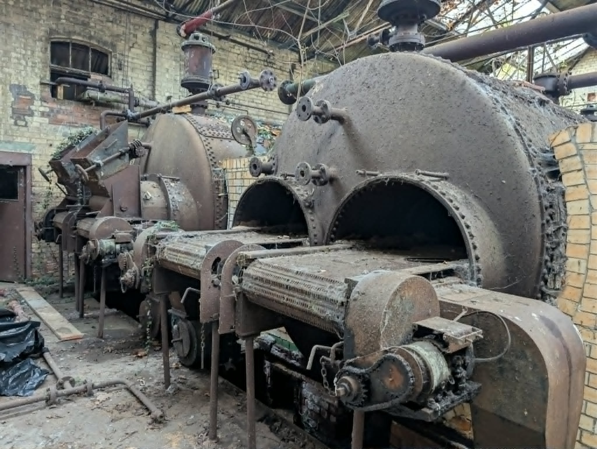 Old, abandoned industrial machinery covered in dirt and rust, situated outdoors near a brick building with a window, surrounded by debris and overgrown plants.