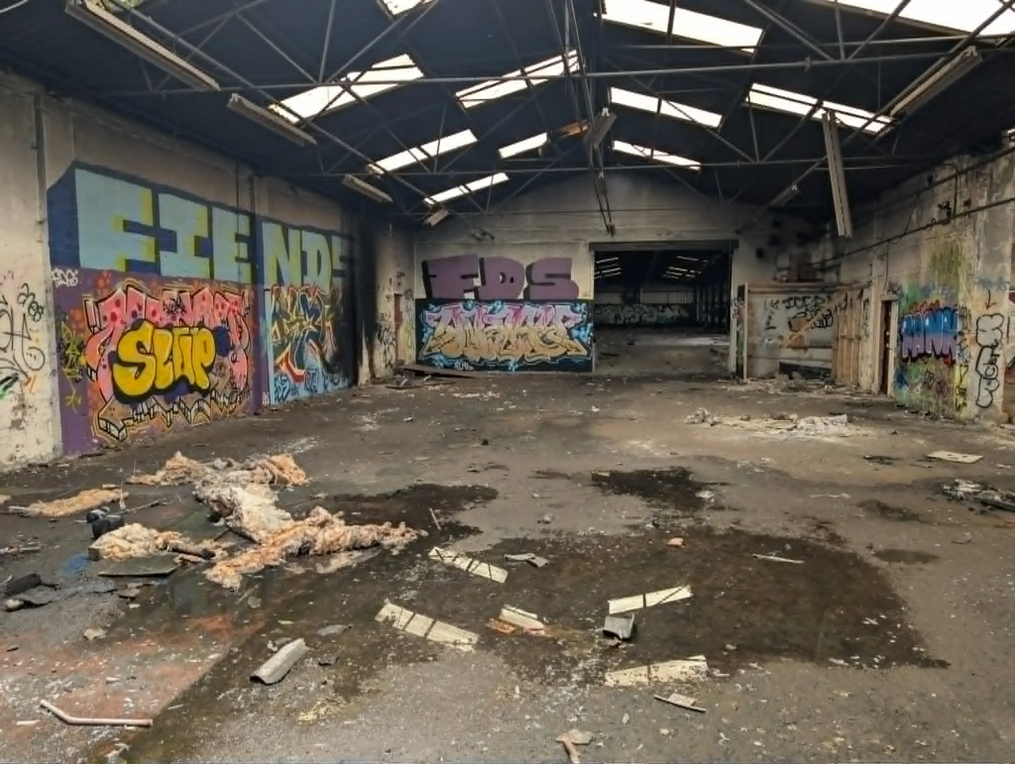 An abandoned building interior with graffiti-covered walls, a damaged metal roof with broken panels, and debris scattered on the ground.