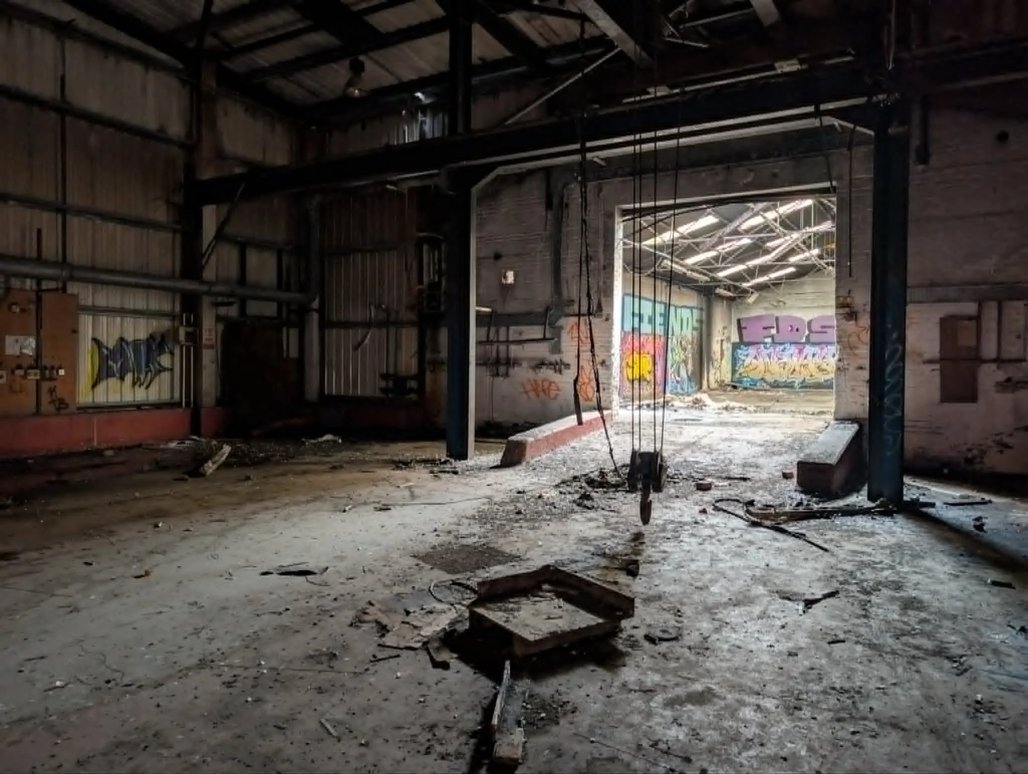An abandoned warehouse with a dirt floor, peeling walls, and graffiti. The ceiling has exposed metal beams and pipes, with some hanging cords. Natural light comes through a large opening showing more graffiti on the distant wall.