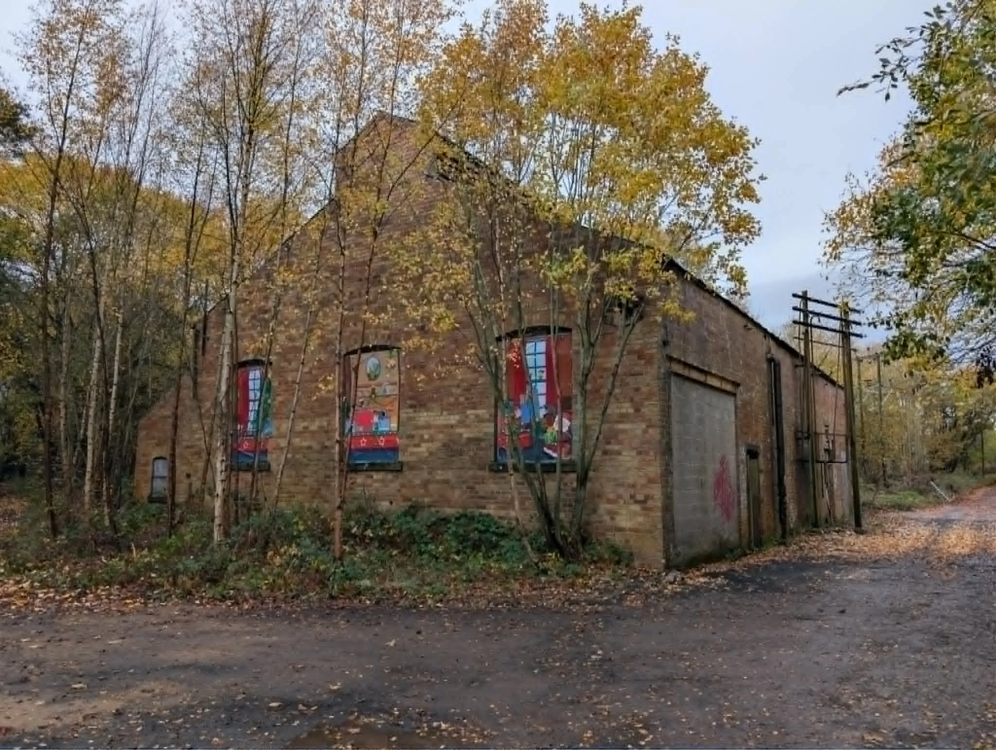 A brick building with colorful banners hanging from the windows, surrounded by trees with autumn leaves, and a dirt road in the foreground.