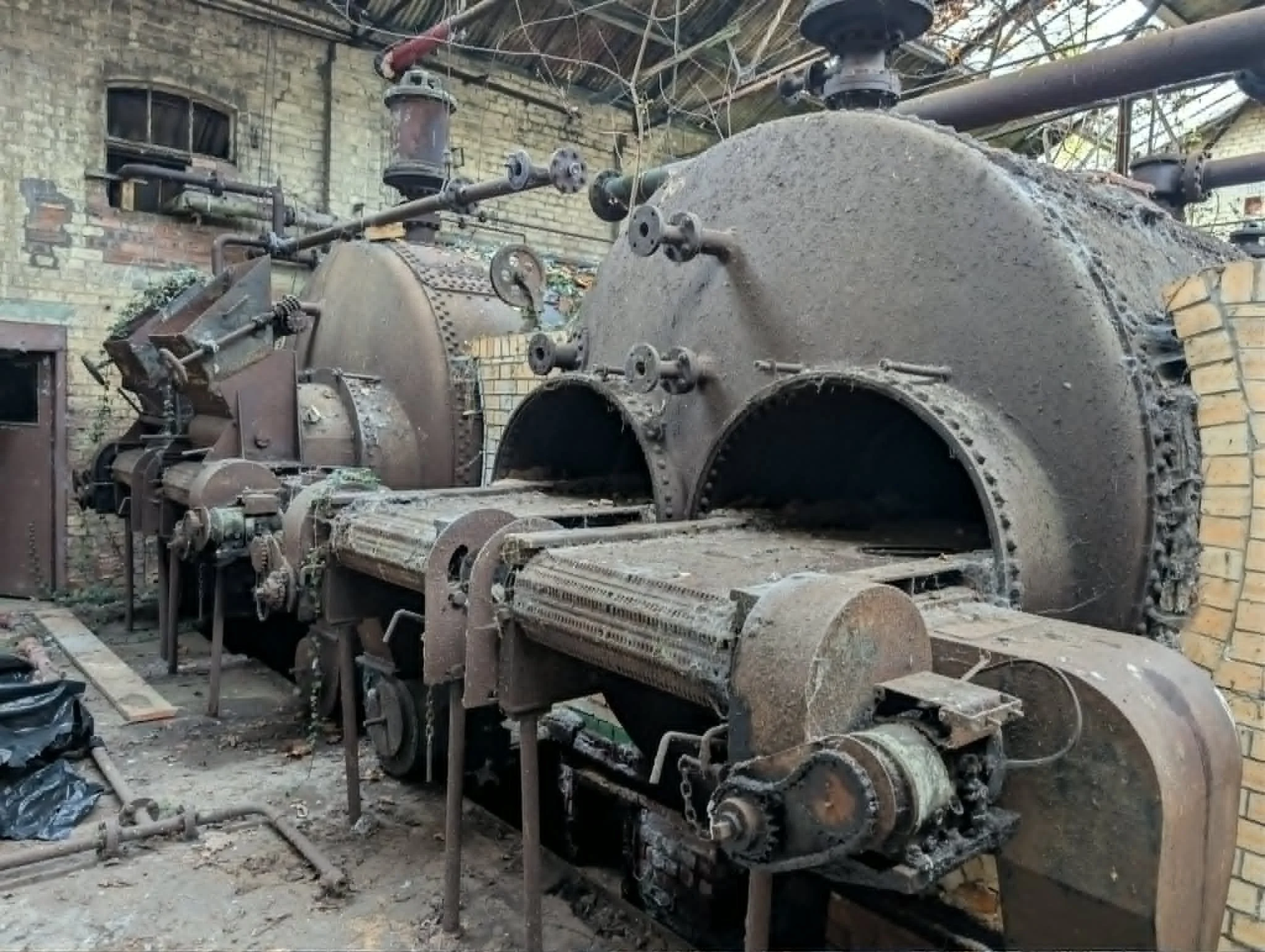 Old, abandoned industrial machine covered in dust and rust in a workshop with brick walls and a corrugated metal roof.