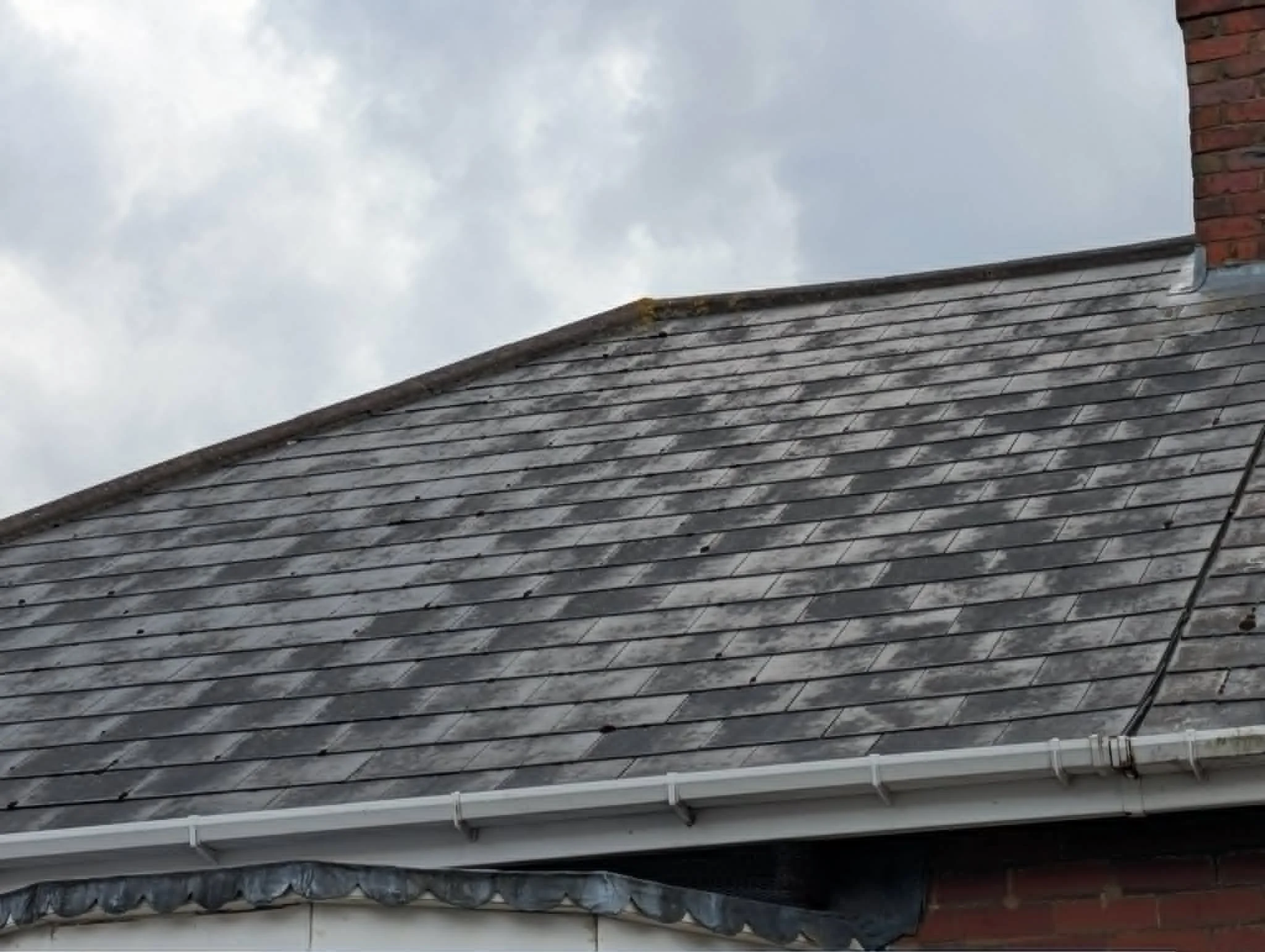 View of a residential house roof with multiple layers of shingles, a brick chimney at the top right, and part of the white gutter system at the bottom; overcast sky background.