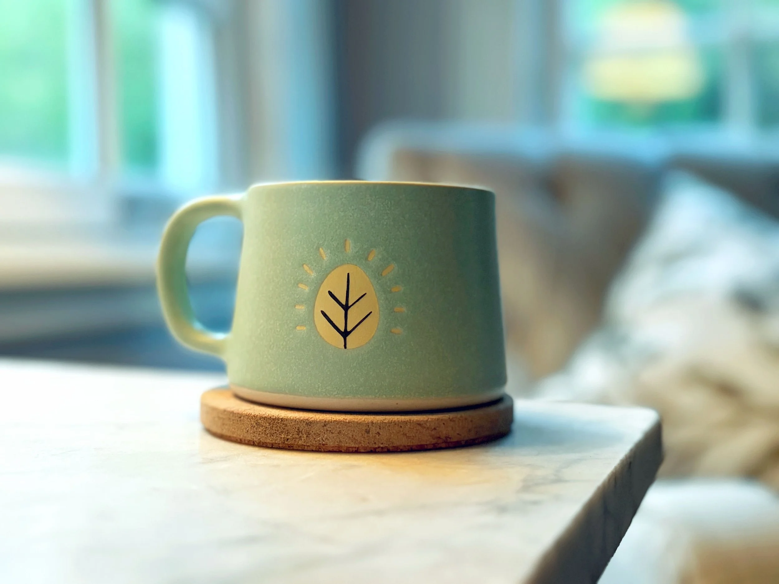 A teal ceramic coffee mug featuring a yellow leaf, which is Sugar Leaf's iconic brand mark. The mug is sitting on a wooden coaster on a light-colored surface in a cozy, homey setting.