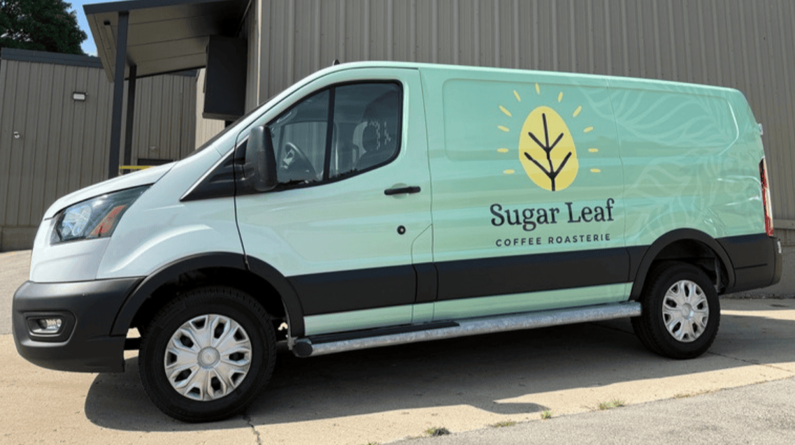 The Sugar Leaf delivery van with the logo for Sugar Leaf Coffee Roasterie on the side, with teal coloring at the back of the van that fades to white at the front. The van is parked outside a building.