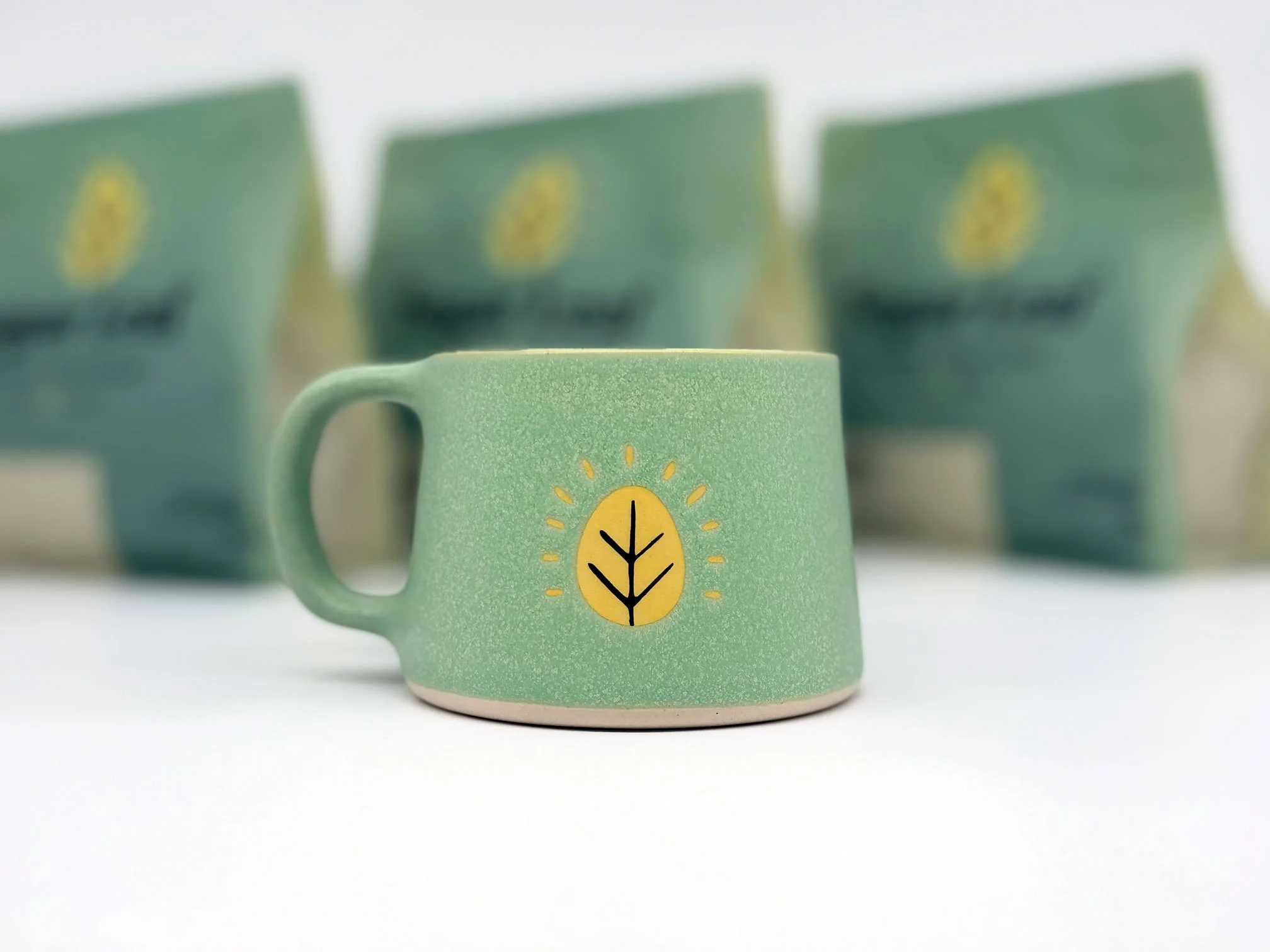 Sugar Leaf ceramic coffee mug, teal with the brand's yellow leaf mark with coffee bags blurred in the background