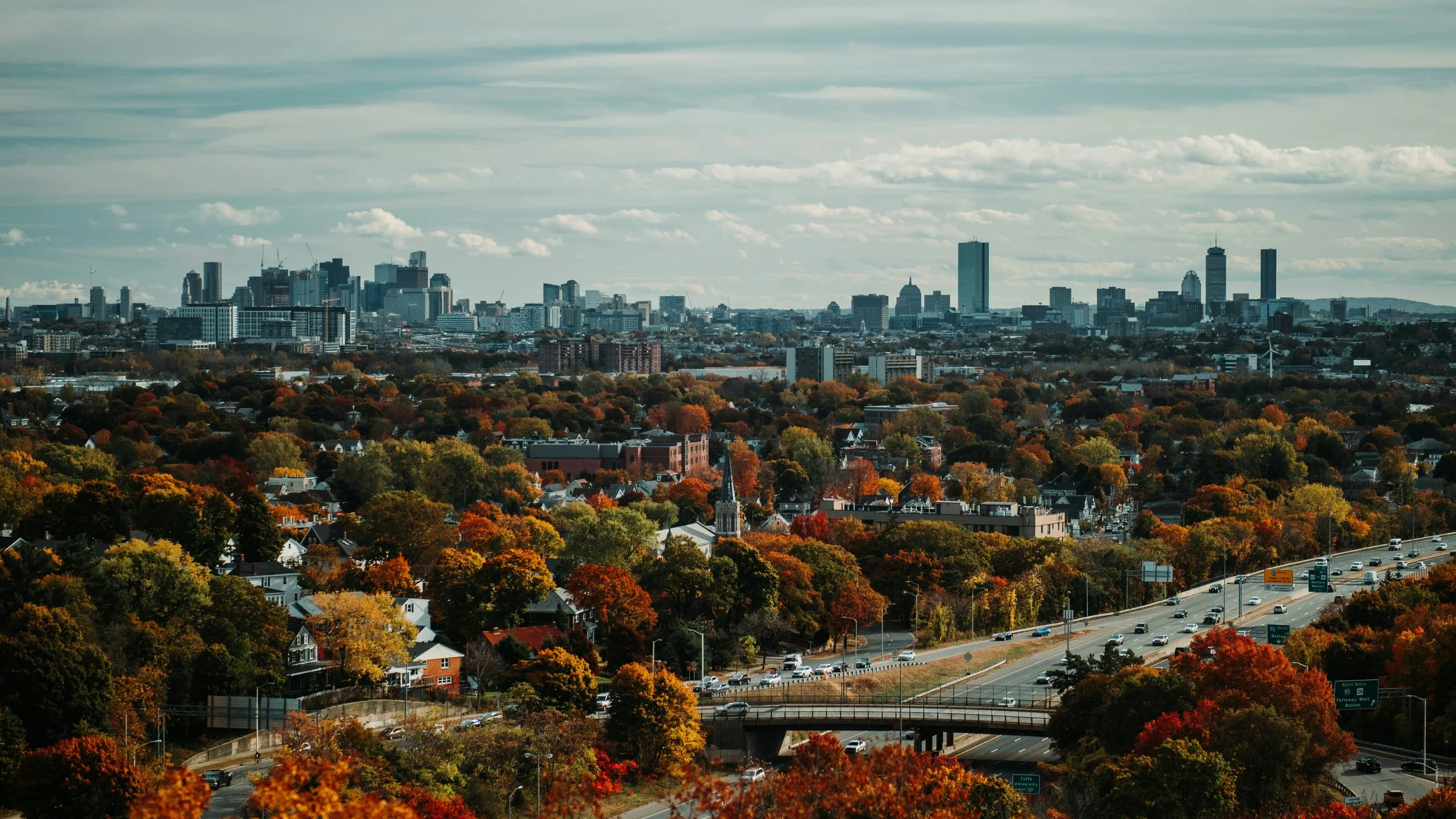 City skyline with tall buildings in the distance and colorful autumn trees in the foreground, with a highway passing through.