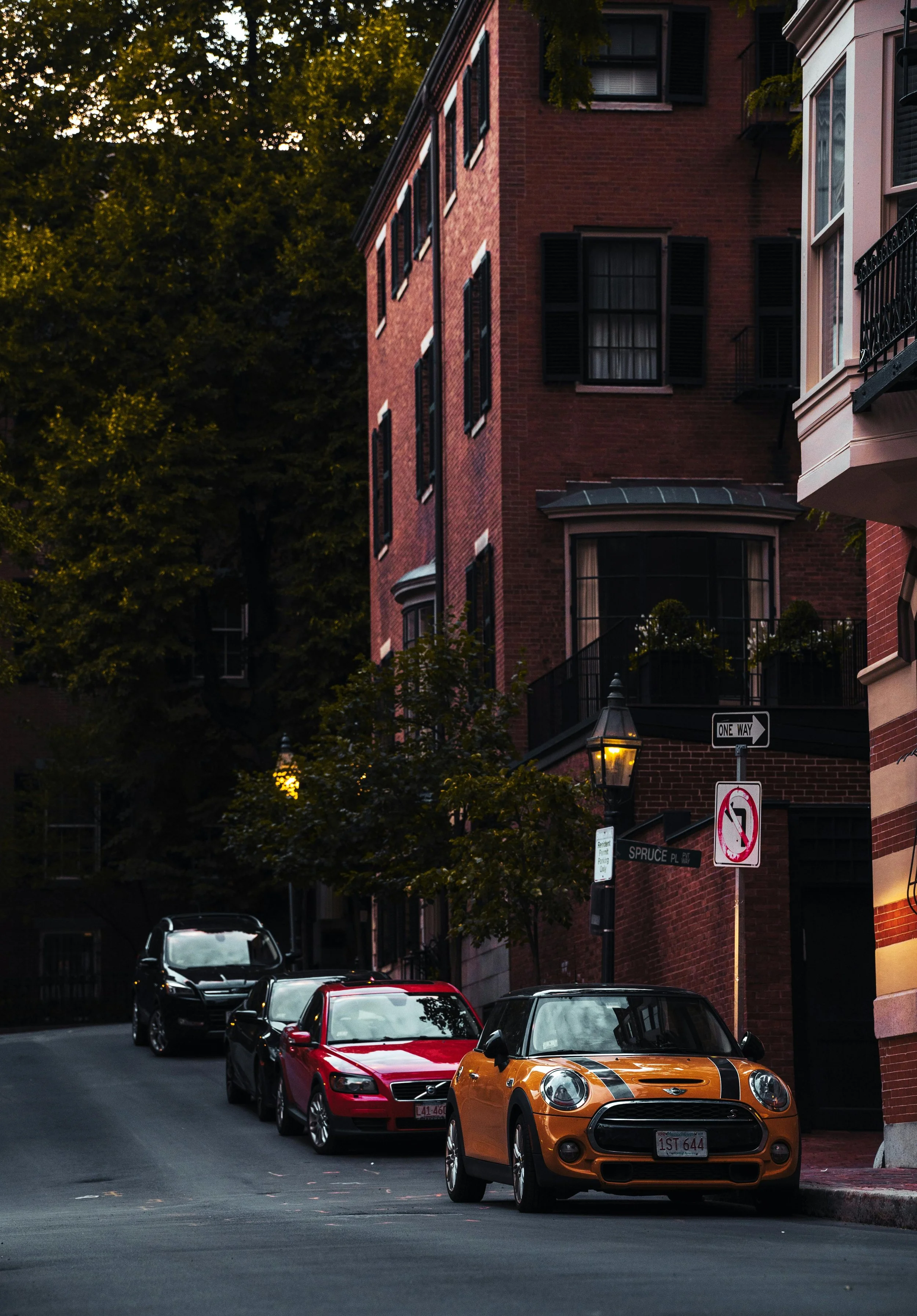 A city street scene during evening with several parked cars including an orange Mini Cooper, a red car, and a black car, along with brick and pastel-colored buildings, street signs including a one-way sign and a no U-turn sign, and trees lining the street.