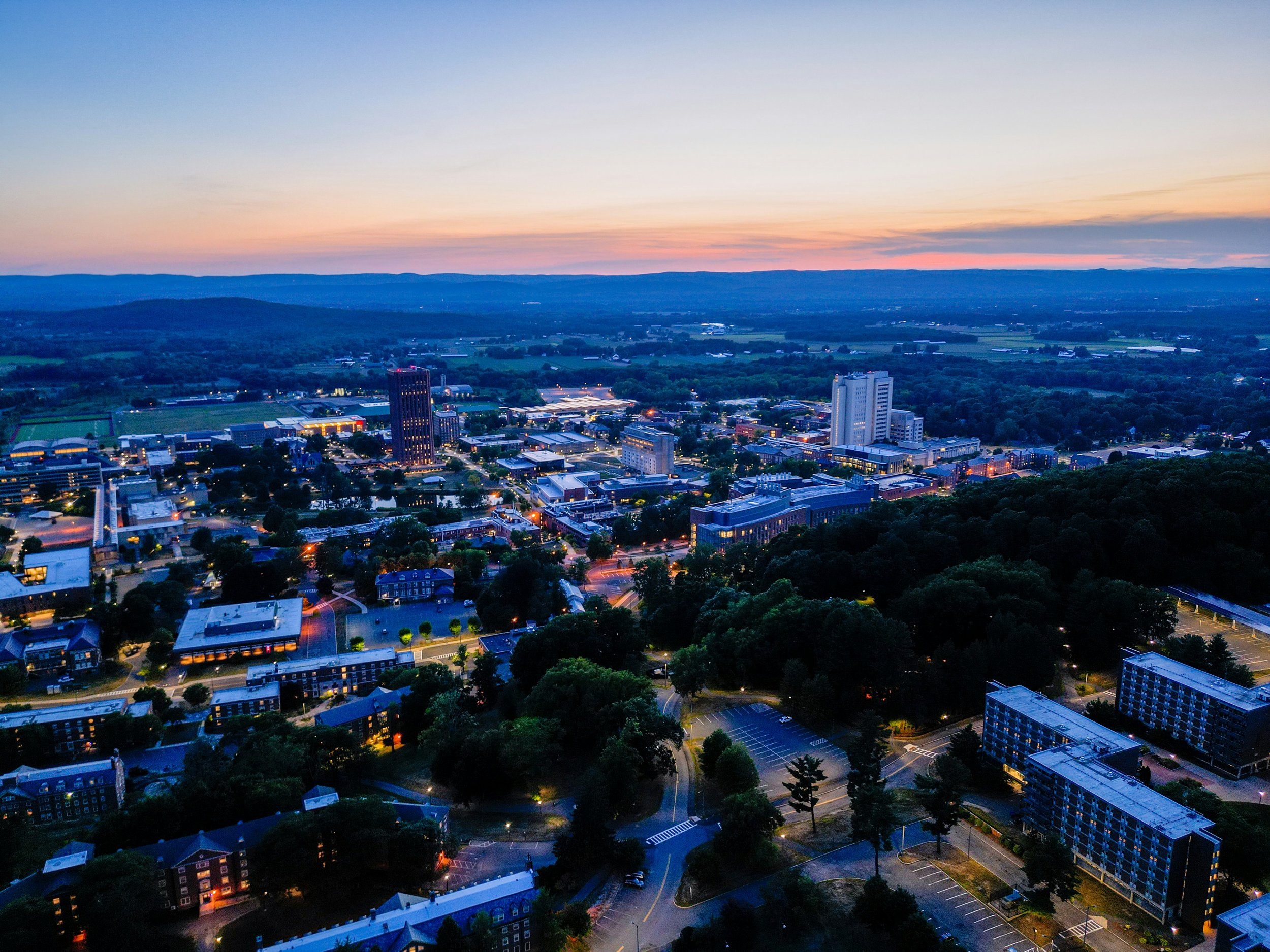 Aerial view of a city at dusk with high-rise buildings, streets, and green spaces.