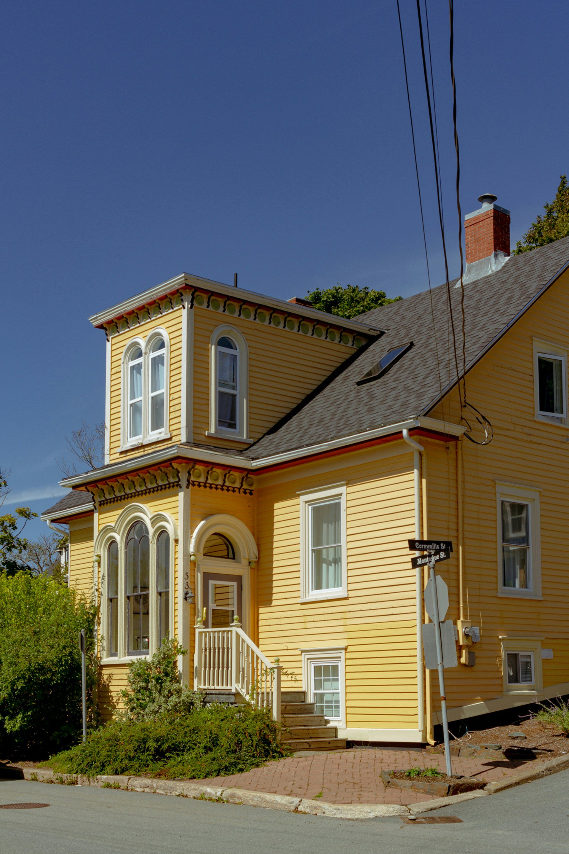 Yellow multi-story house with Victorian architectural details, white trim, and an arched front window, situated at the corner of two streets with street signs reading Cornallies St. and Montague St.
