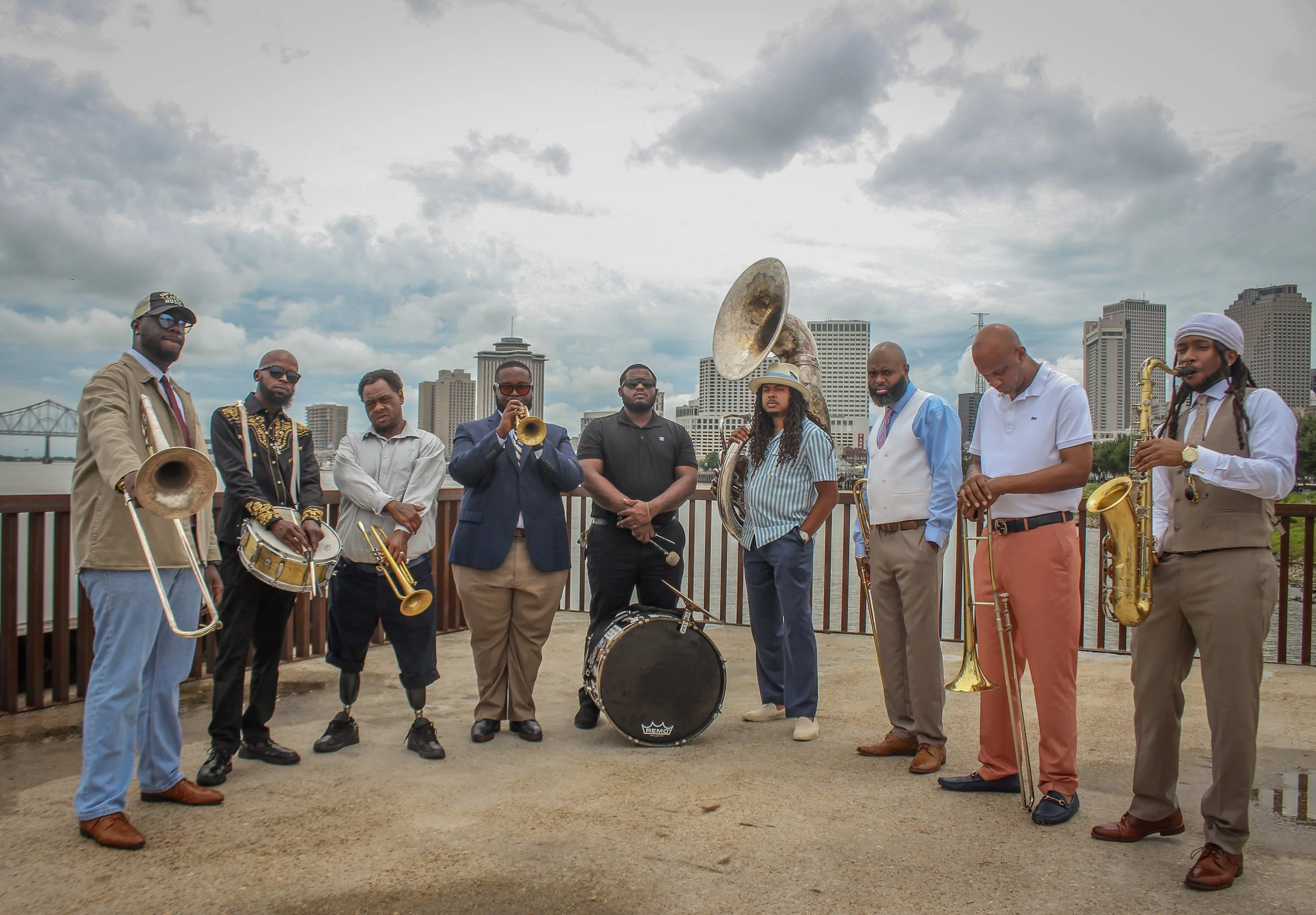 A group of nine musicians standing outdoors in a cityscape, holding various musical instruments including a trumpet, trombone, saxophone, and drums, with a cloudy sky and tall buildings in the background.
