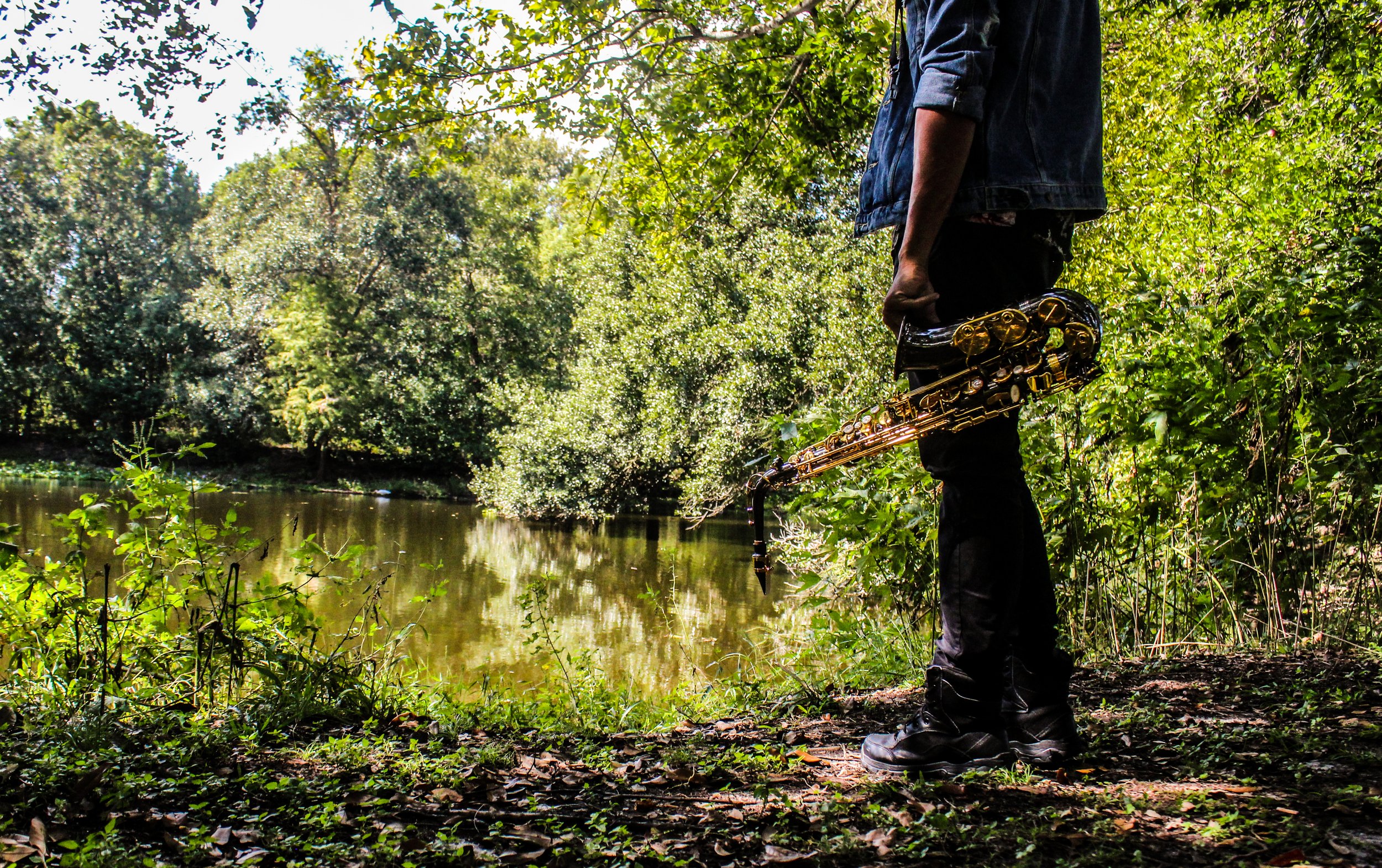 A person standing by a body of water surrounded by trees, holding a saxophone in their right hand, with only their torso and legs visible in the image.