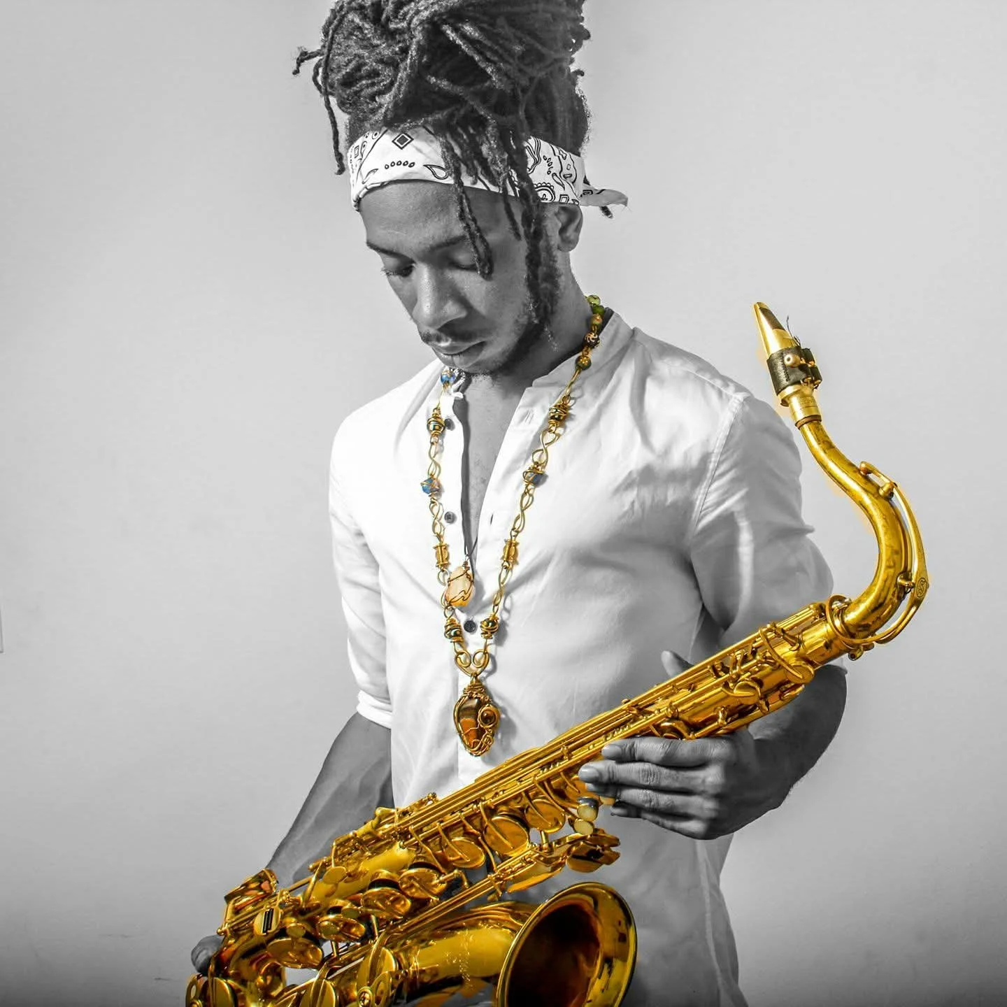 A man with dreadlocks, wearing a white shirt, a bandana, and gold jewelry, holding a gold saxophone. The image is in black and white except for the saxophone and jewelry, which are in color.