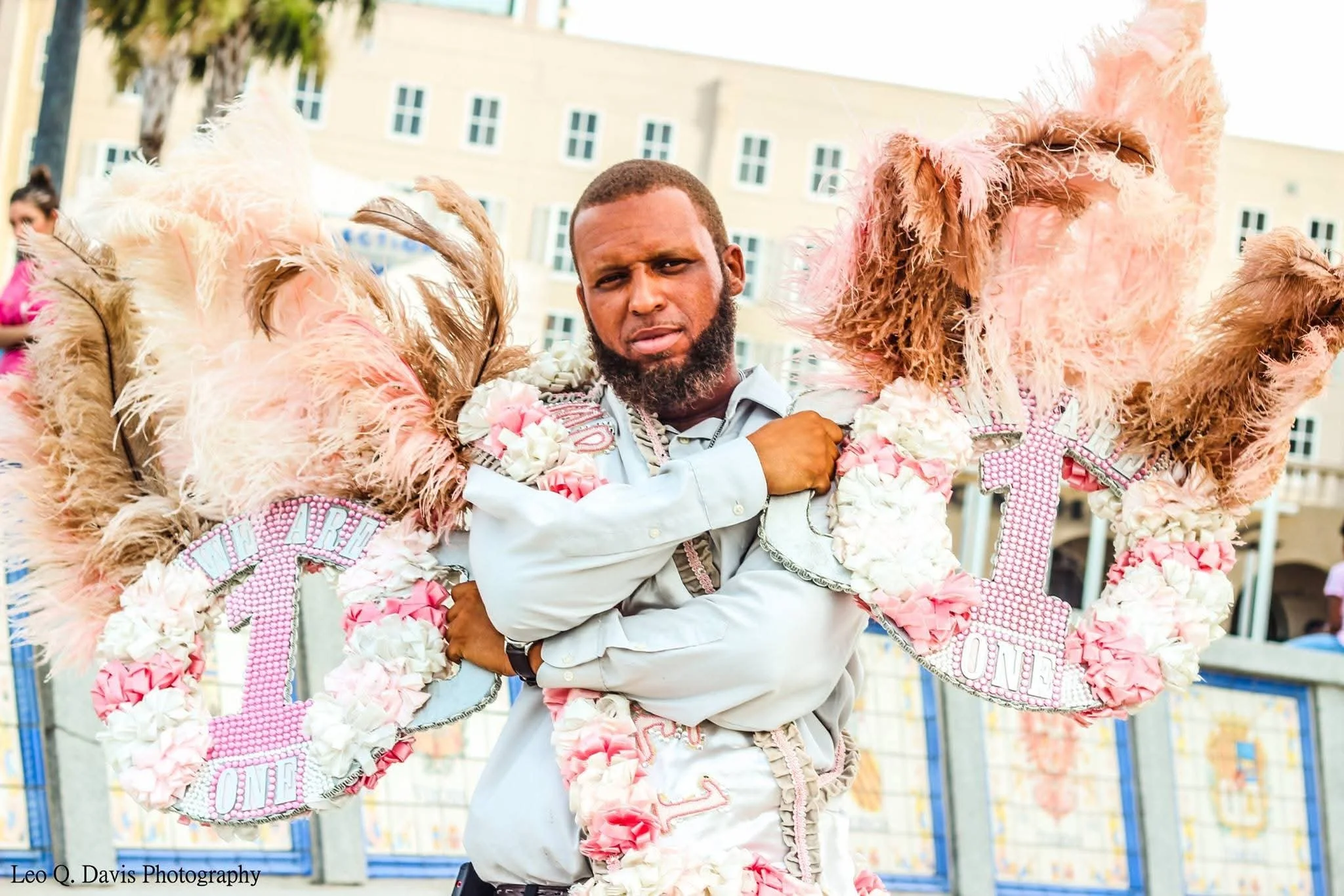 A man with a beard wearing a white shirt standing among pink and white feathered and beaded decorations, with a building and people in the background.