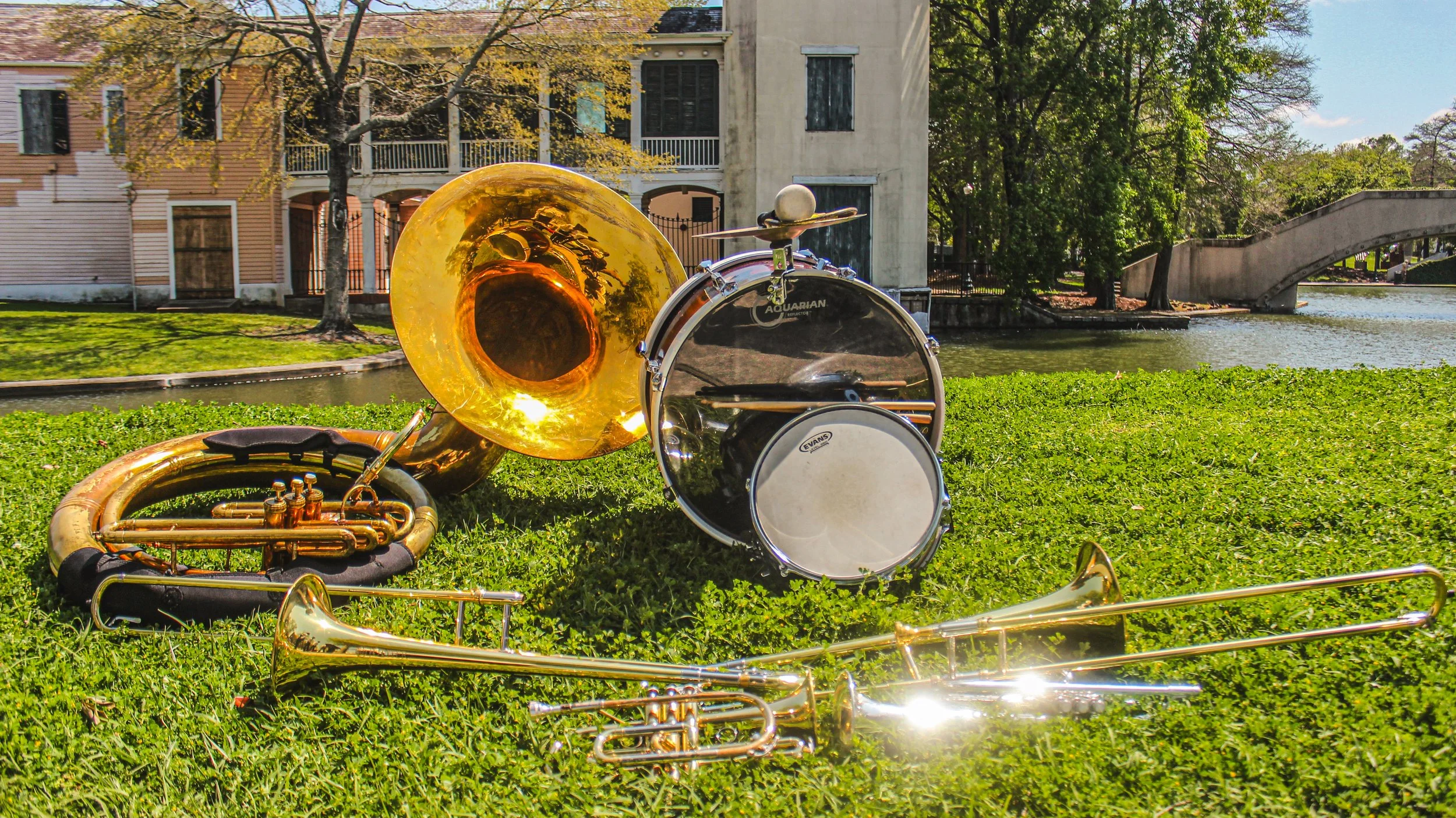 A brass tuba and a snare drum with a drumstick rest on a grassy area near a pond, with trees, a bridge, and residential buildings in the background.
