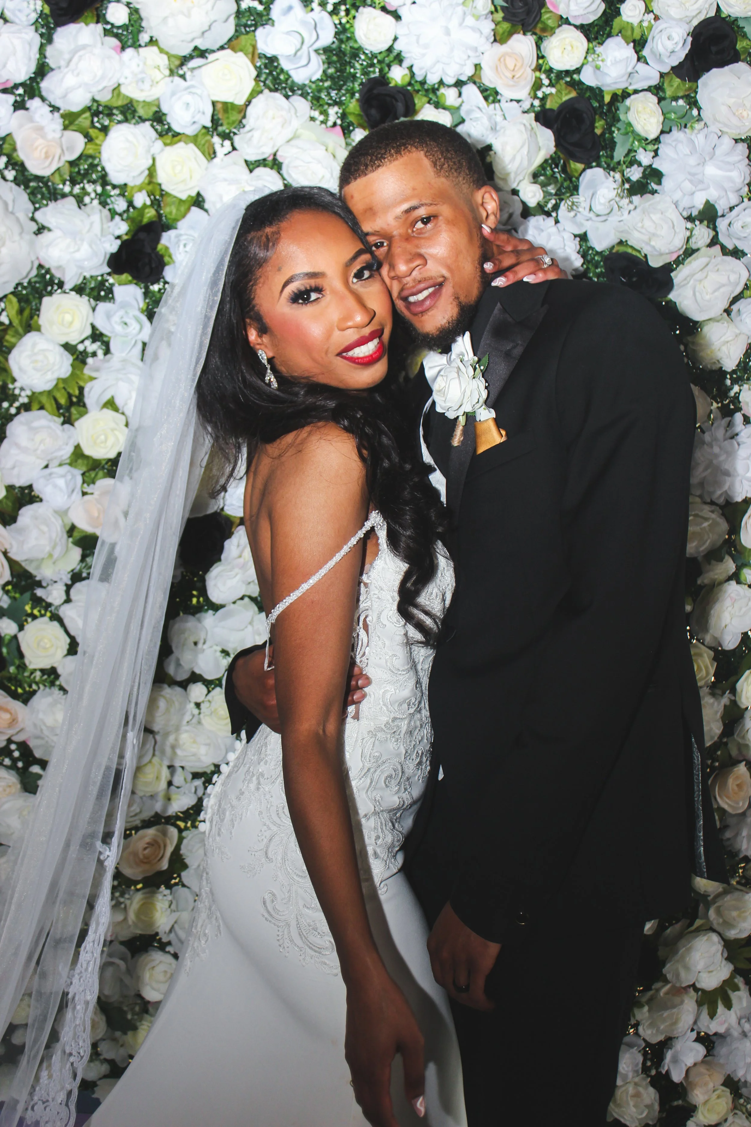 A bride and groom embracing in front of a wall of white and black roses, dressed in wedding attire, with the bride wearing a white wedding gown and veil, and the groom wearing a black tuxedo.