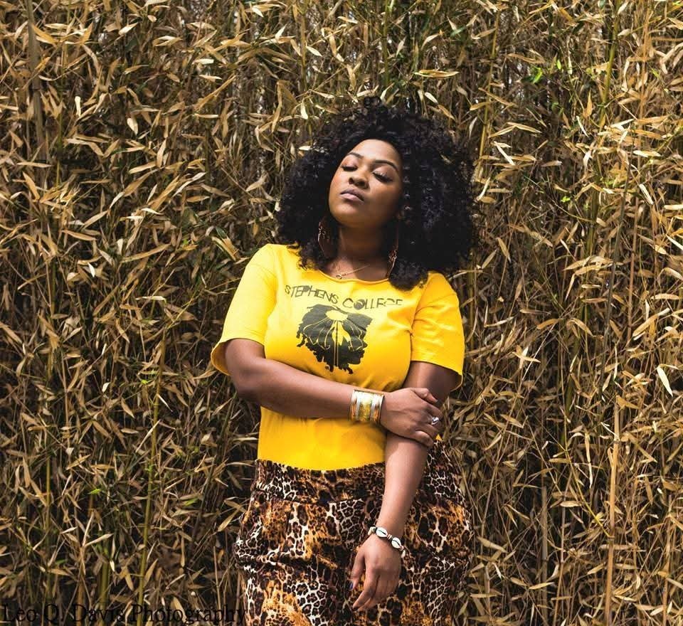 A woman with curly black hair stands in front of a dense background of dried grass or reeds. She is wearing a yellow T-shirt with black text and an image, paired with leopard print pants. She has jewelry, including bracelets and a necklace, and has a confident expression with her eyes closed.
