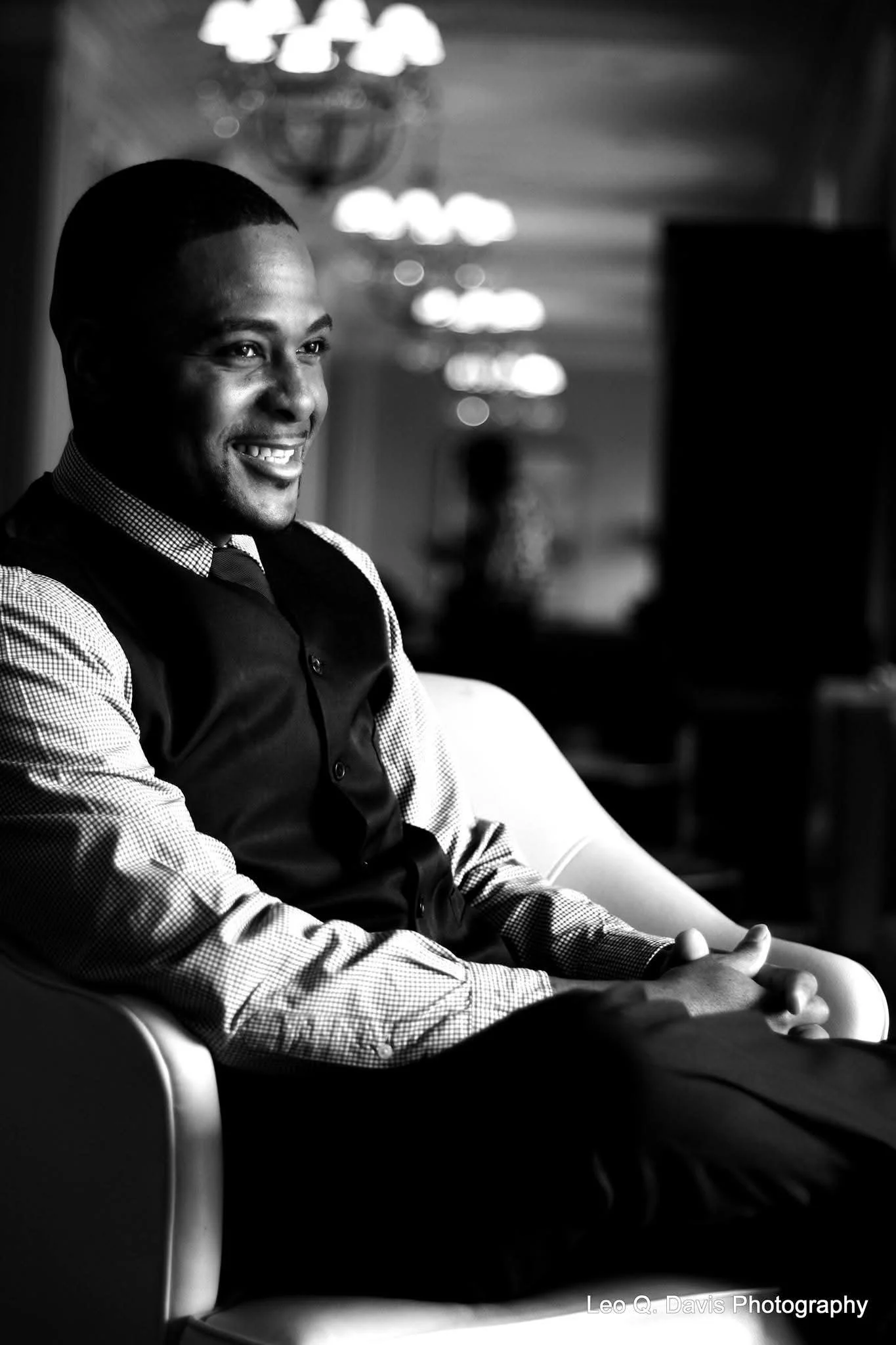A smiling man wearing a checkered shirt and vest sitting in a chair in a room with blurred lights and decor in the background, black and white photography.