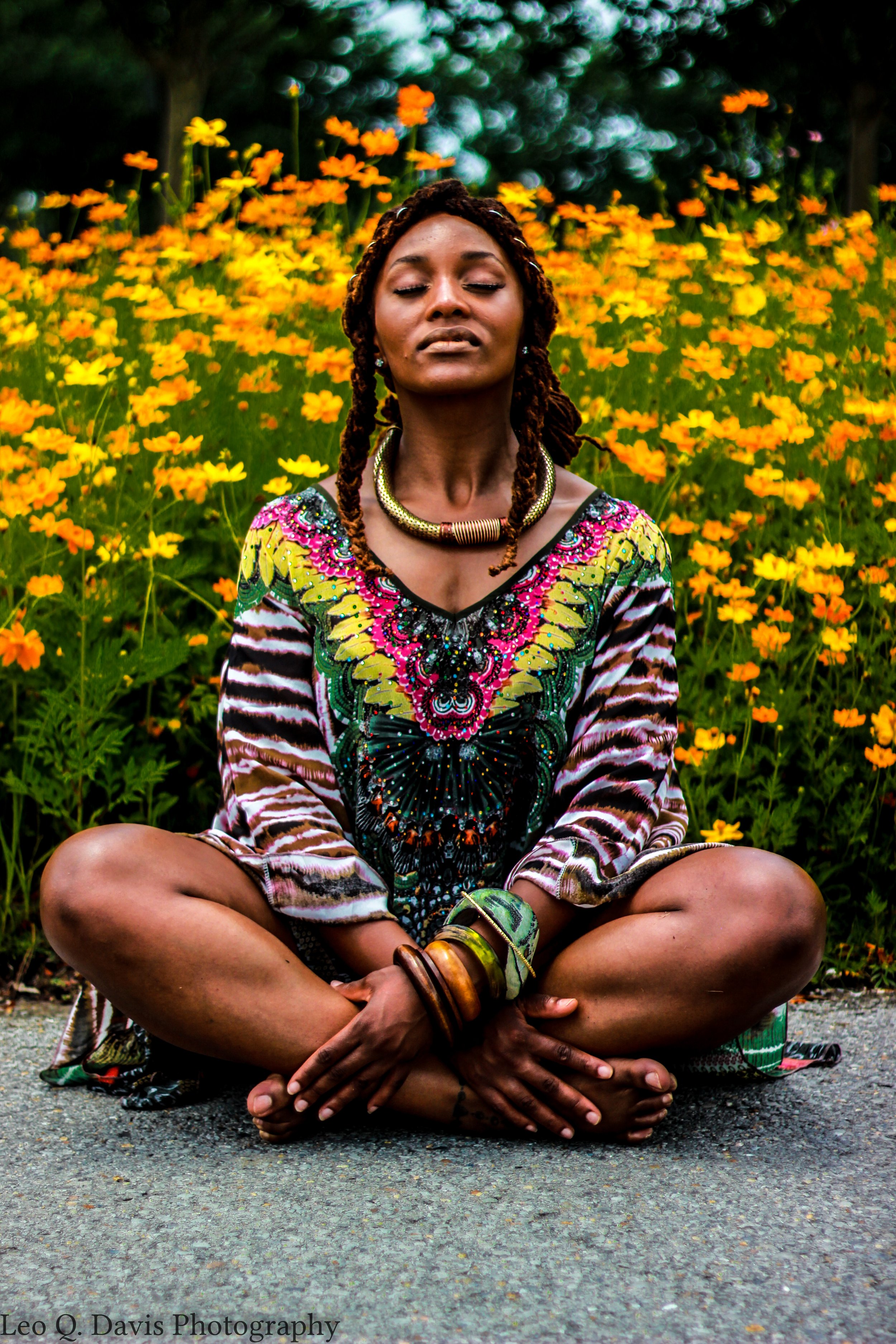 A woman sitting cross-legged on the ground with her eyes closed, surrounded by a field of yellow and orange flowers, wearing colorful clothing and jewelry.