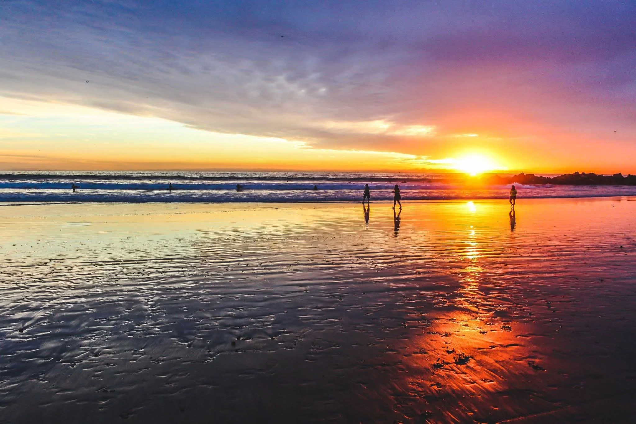 Sunset over the ocean with a colorful sky, reflecting on wet sand, with people walking along the beach and seagulls flying overhead.