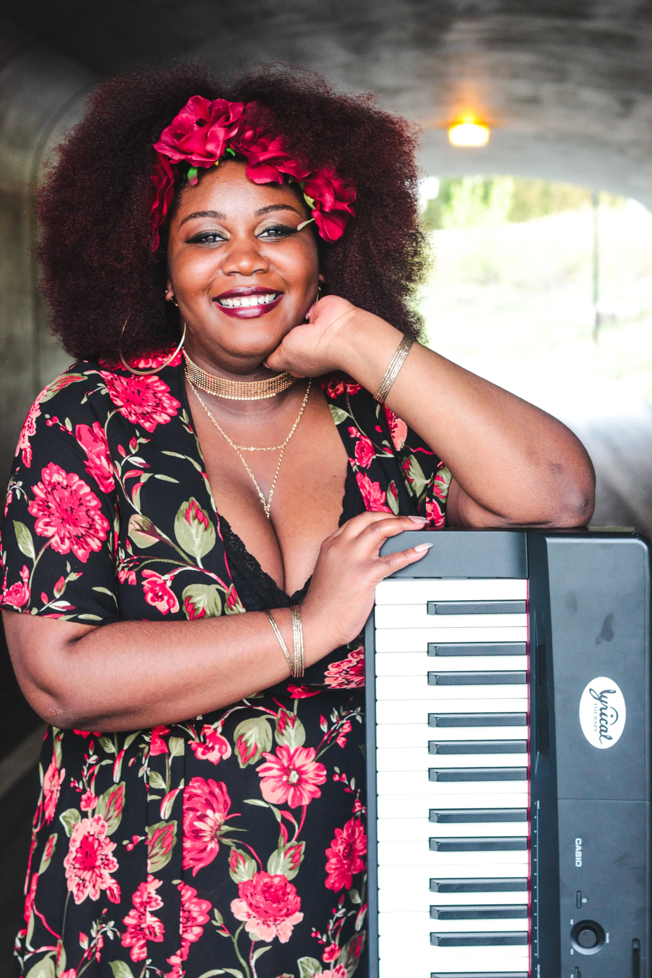 A smiling woman with curly hair wearing a red floral headband and floral dress, holding a Casio musical keyboard and sitting under a bridge or tunnel.