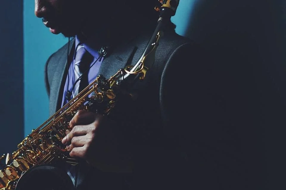 A person in a suit playing a gold saxophone with a blue background.