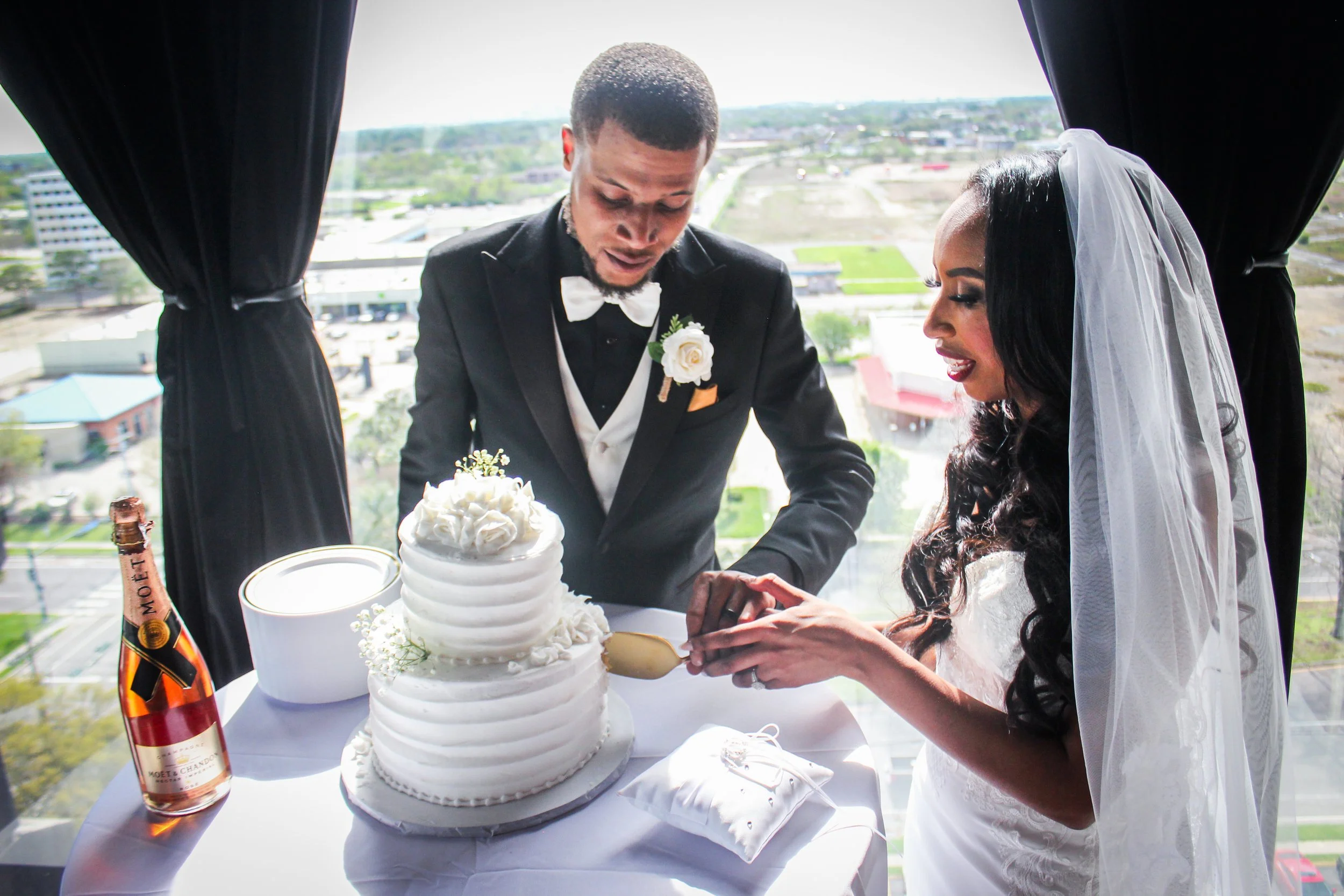 A bride and groom in wedding attire are cutting a white wedding cake together during their wedding celebration. The groom is wearing a black tuxedo with a white bow tie and boutonniere, the bride is dressed in a lace wedding gown with a veil. They ar