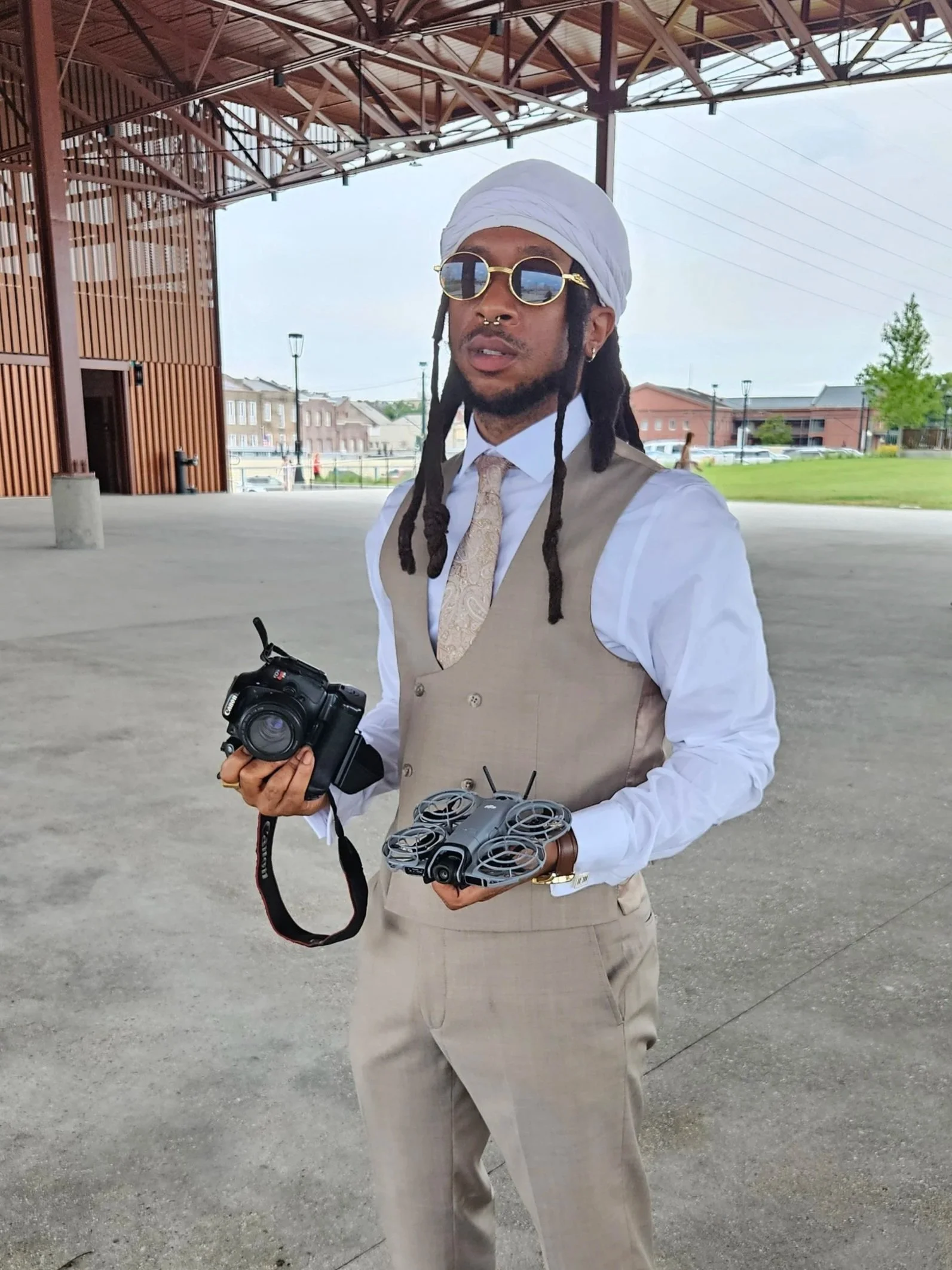 A man dressed in a beige suit with a white shirt and beige vest, wearing sunglasses, a white head wrap, and jewelry, holding a camera and drone controllers under a large open pavilion with wooden support beams, in a suburban area with houses, trees, and cars in the background.