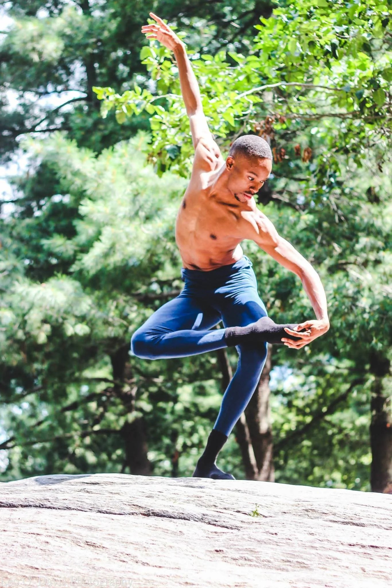 A young man performing a yoga pose outdoors on a large rock, surrounded by green trees.