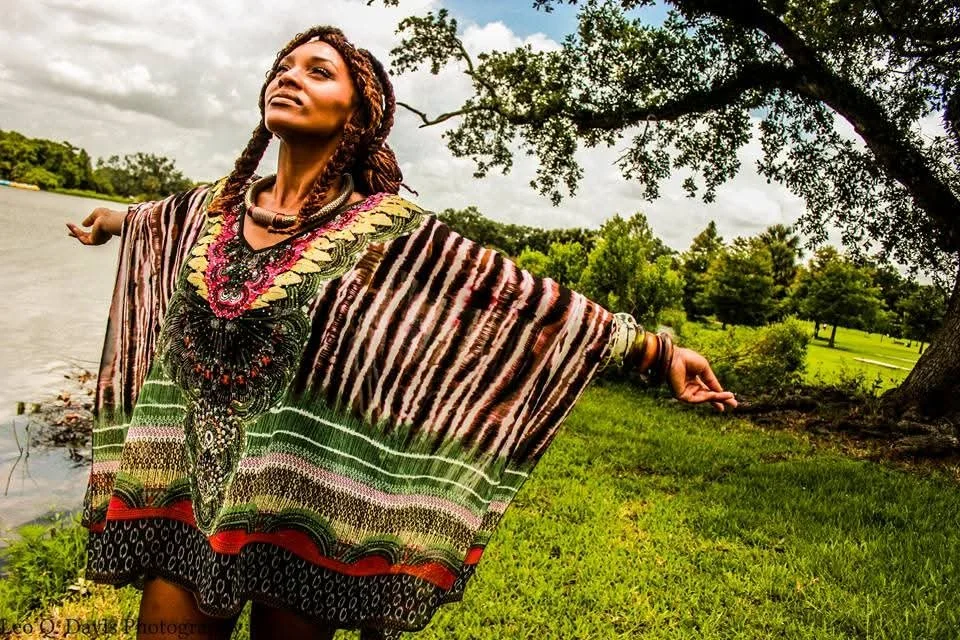 A woman wearing a colorful, patterned dress with her arms outstretched outdoors near a body of water, with trees and grass in the background.