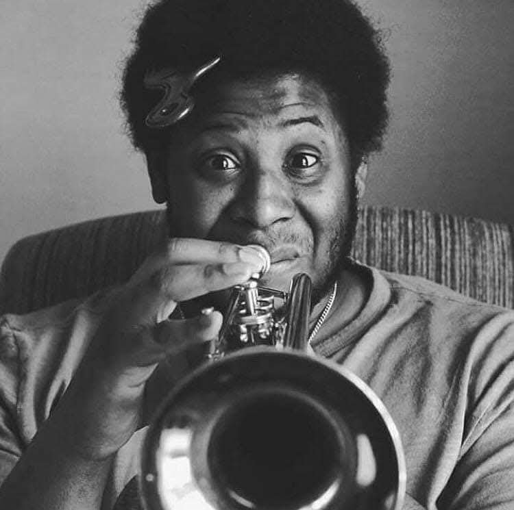 A man with curly hair and a clip in his hair, playing a trumpet and smiling at the camera in a black and white photo.