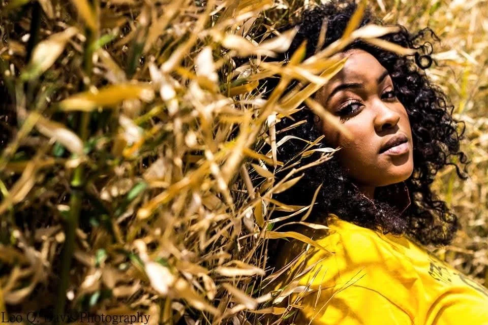 A woman with curly black hair wearing a yellow shirt, lying in a field of tall, dry grass or wheat, with a serious expression and looking at the camera.