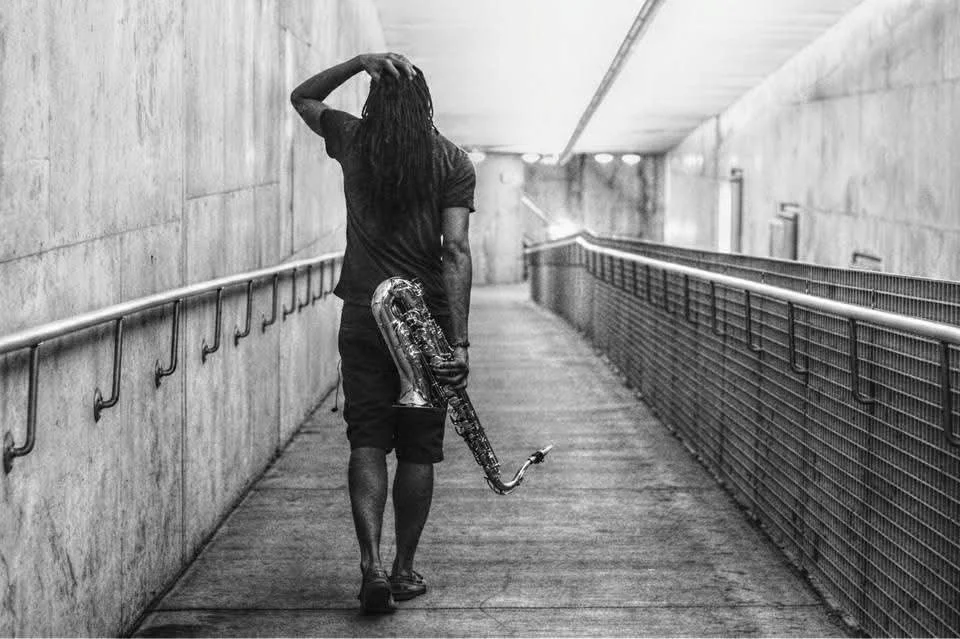 A person walking through an underground tunnel with concrete walls, holding a saxophone and resting their hand on their head.