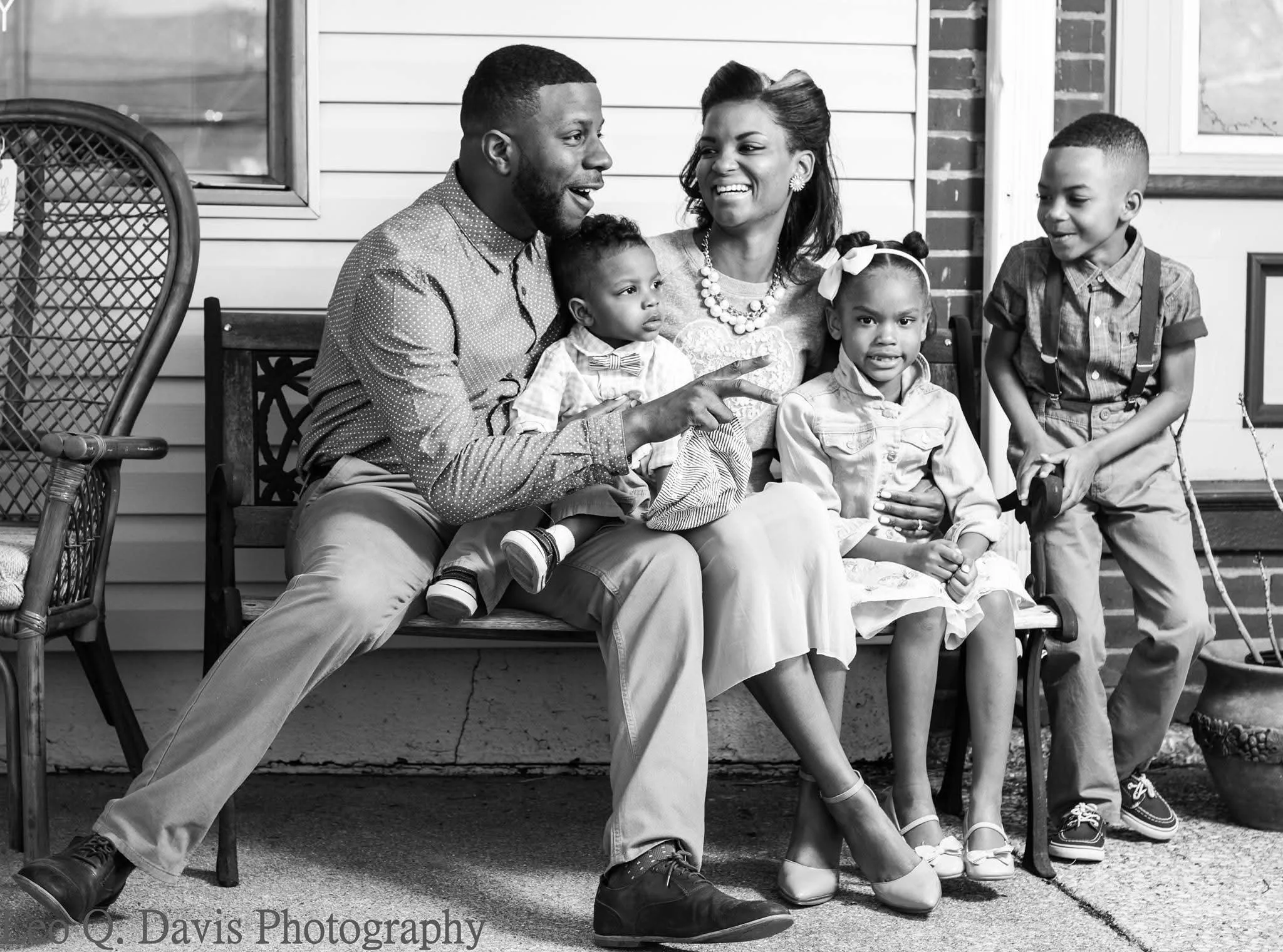 A black and white photo of a happy family sitting on a porch. The man and woman are smiling at each other, with the man making a peace sign. Two young children, a girl and a boy, sit with them. The girl is wearing a dress and has her hair in pigtails