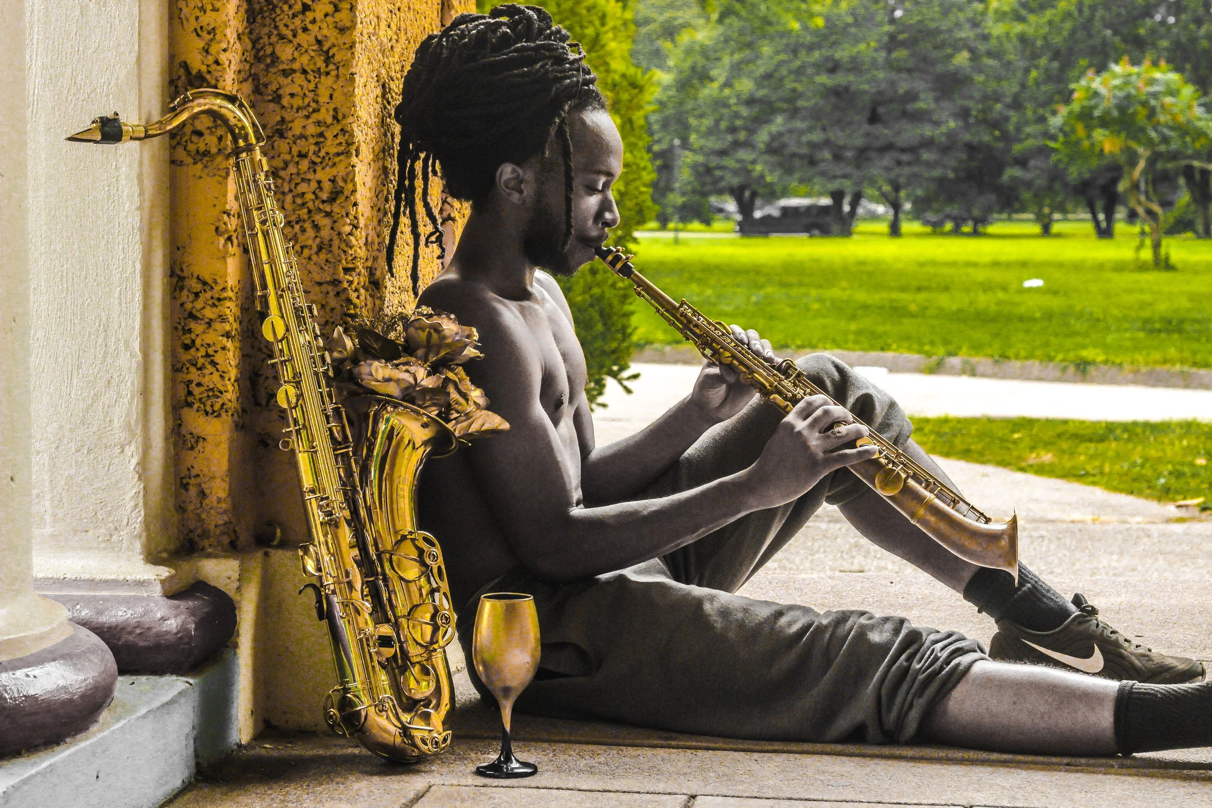 A shirtless man with long dreadlocks sitting on the ground, playing a saxophone, with a saxophone and a drink nearby, against a yellow textured wall in a park with green trees in the background.