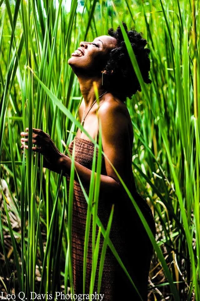 A woman standing in a lush green field of tall grass or reeds, smiling with her eyes closed, wearing a strapless dress.