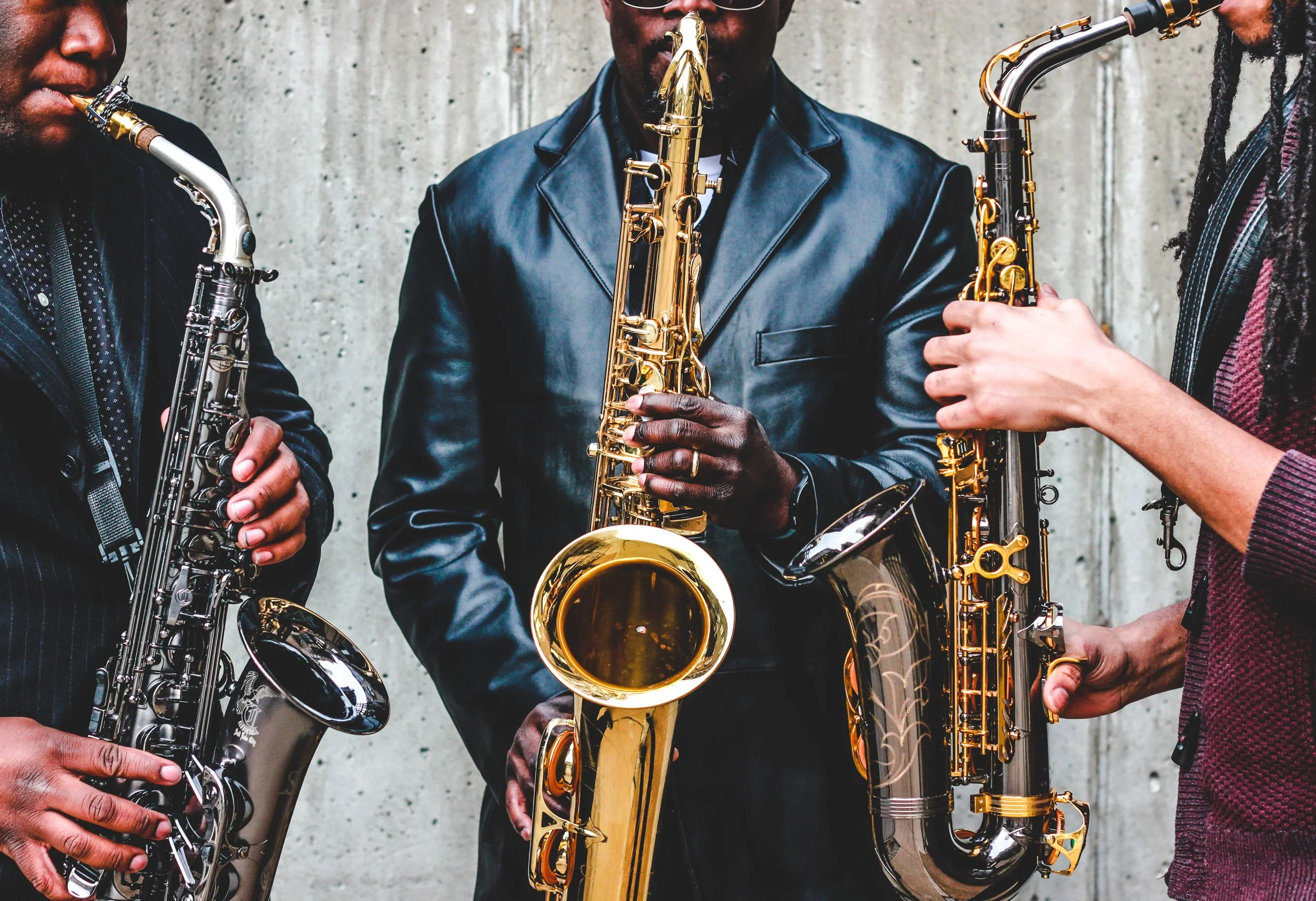 Close-up of three musicians playing saxophones, with a concrete wall background. The musicians are wearing dark clothing, and the photo captures their instruments and hands.