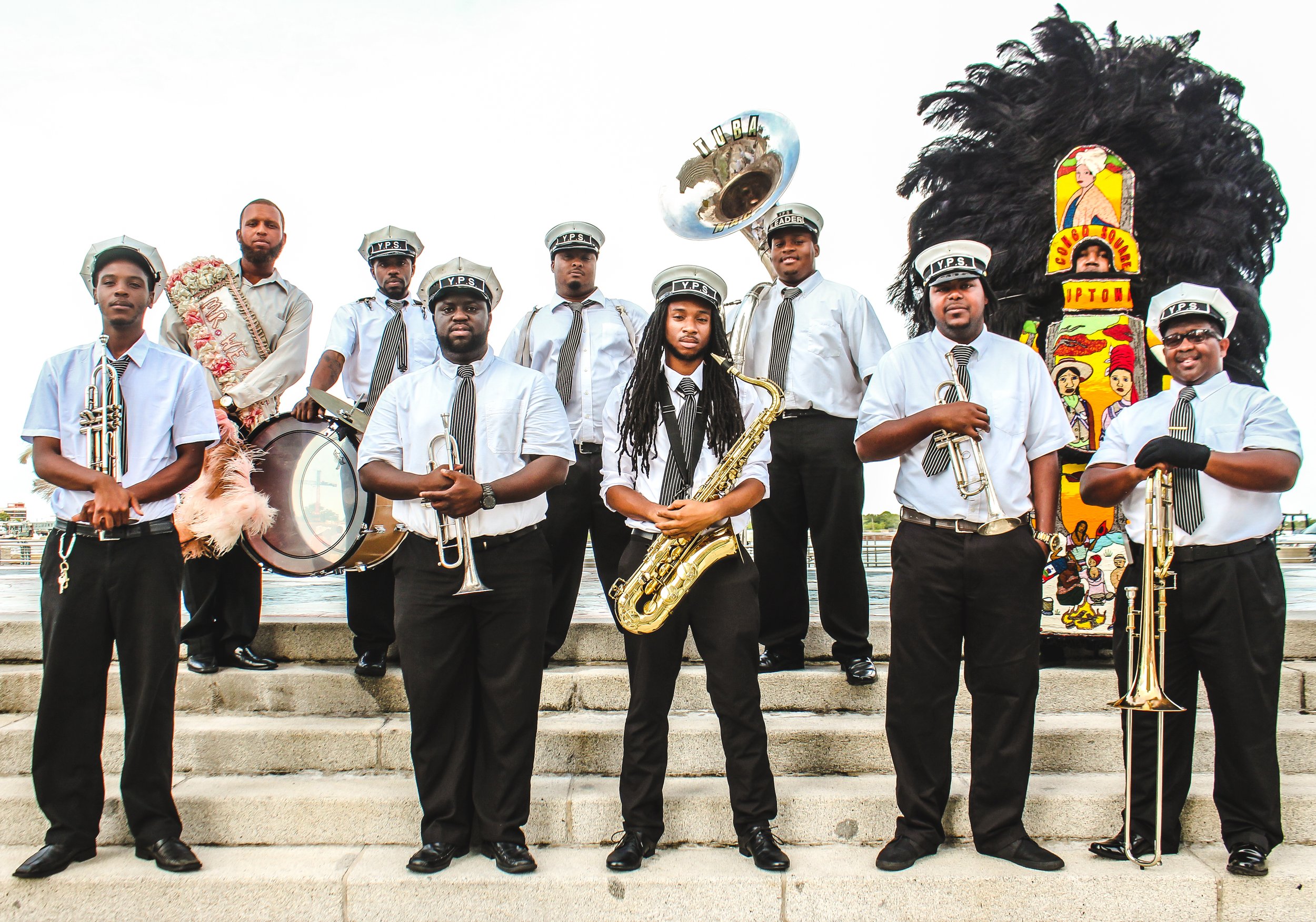A group of musicians in uniform standing outdoors, with some holding instruments such as saxophone and trumpet. There is a large colorful totem pole on the right side of the image and a reflective bell with the word "TUBA" on it in the background.