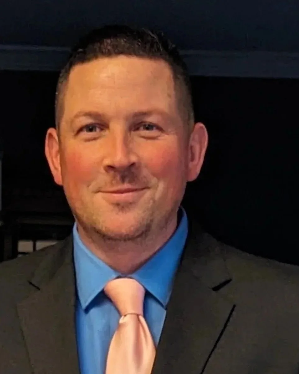 A man in a suit smiling, wearing a blue shirt and peach-colored tie.