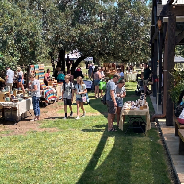 People browsing outdoor craft market with tables displaying handmade items, shaded by trees on a grassy area.