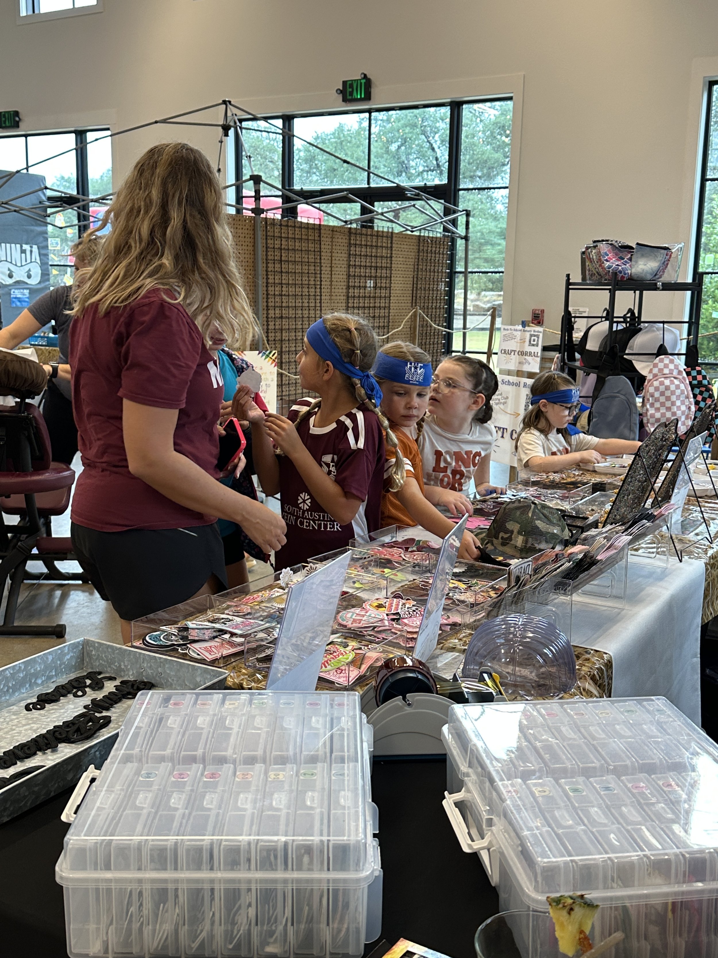 A group of children talking to an adult girl with long blonde hair at a craft fair booth with various craft supplies and jewelry displays.