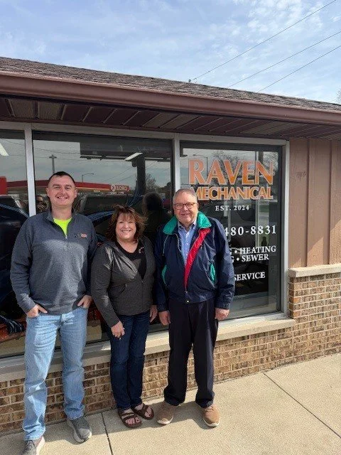 Three people standing outside Raven Mechanical storefront, smiling, with the store's sign visible.