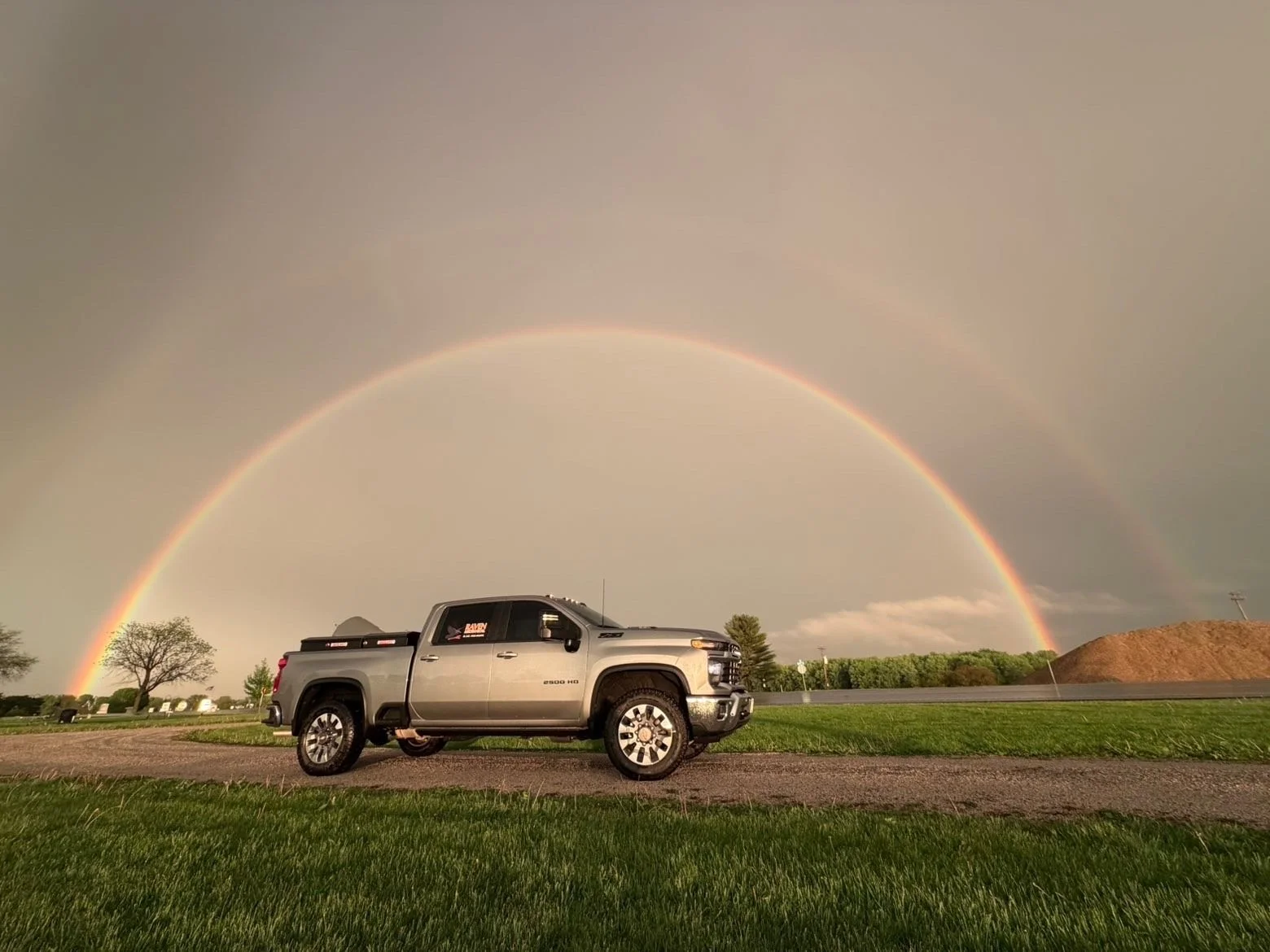 Silver pickup truck parked on a grassy area with a rainbow in a cloudy sky and a few trees and a small hill in the distance.