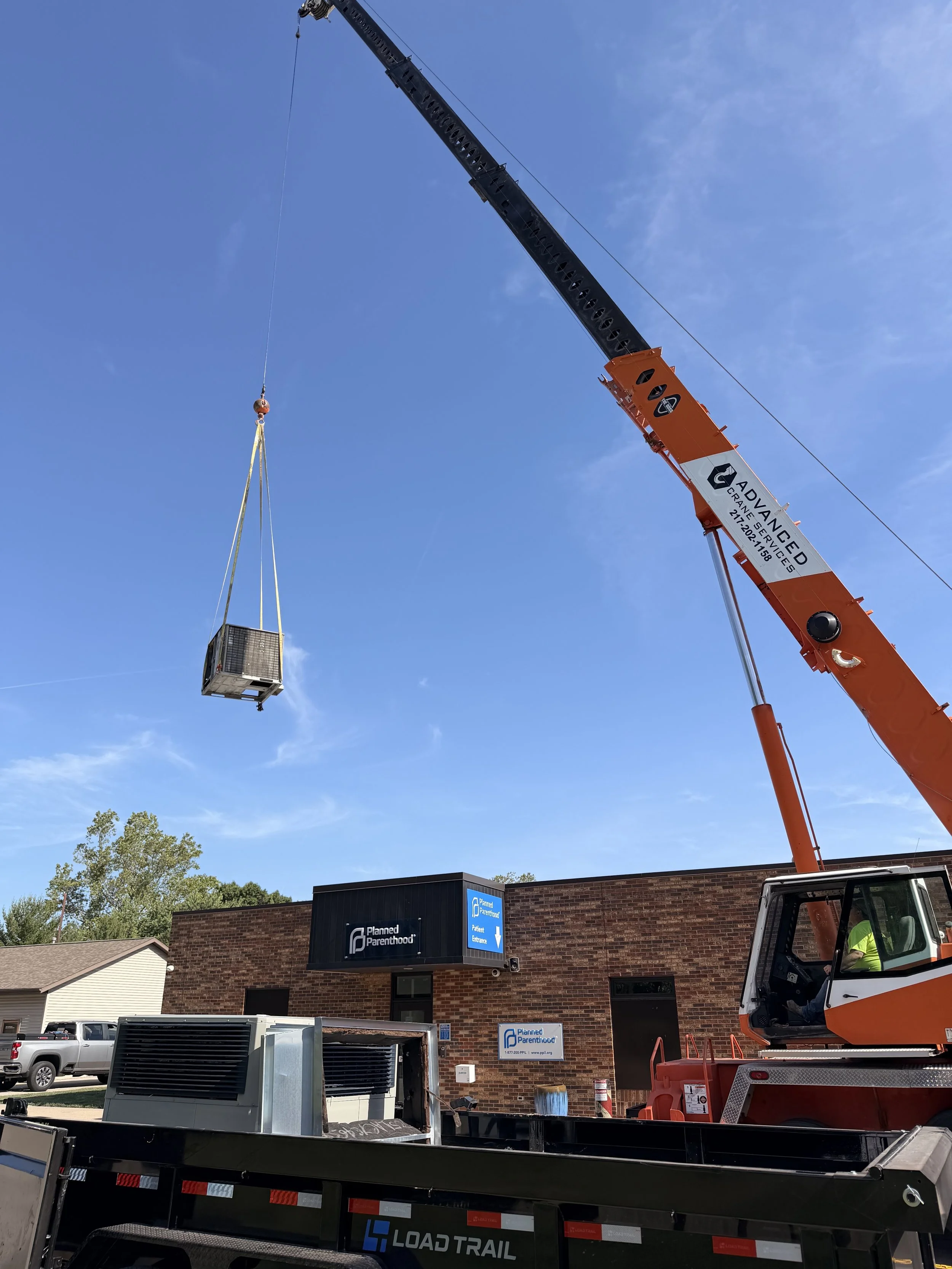 A large orange crane lifting a container with a sign for Planned Parenthood on a brick building. There are parked trucks and trees visible below.