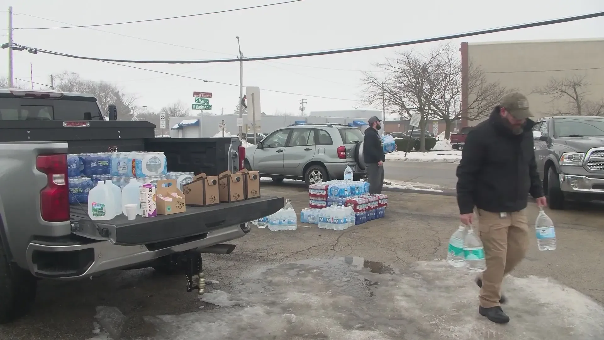 People handing out bottled water and supplies on a snowy street, with vehicles parked nearby, as a makeshift distribution point in cold weather.